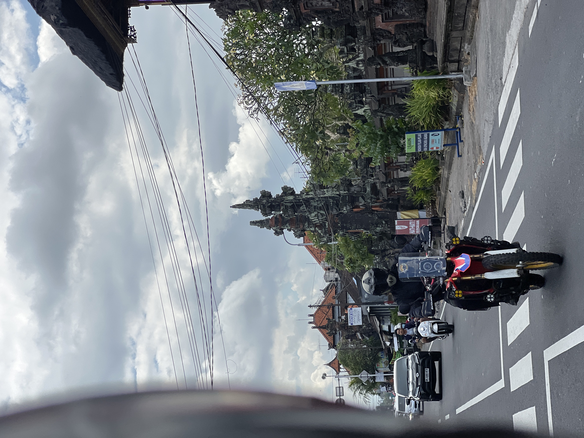This is a street-level, medium-shot perspective from a moving vehicle looking forward on a cloudy day in Abiansemal, Indonesia. The foreground shows a street with white pedestrian crossing lines. In the middle ground, a person in black motorcycle gear and helmet is riding a red and white adventure motorcycle with multiple auxiliary lights. The motorcycle has a transparent windshield with stickers and a holder for a phone or GPS. Behind this motorcycle, a person on a white scooter is visible, followed by a black SUV. The street is bordered by a stone wall adorned with carved sculptures, characteristic of Balinese architecture. Trees with white flowers line the wall. Several signs are posted along the sidewalk, including a blue sign with white text that reads "MOHON PELAN-PELAN" and "ADA UPACARA AGAMA" (Please slow down, there is a religious ceremony). Further down the street, traditional Balinese buildings with orange tiled roofs are visible under the cloudy sky. Power lines are strung overhead across the scene.