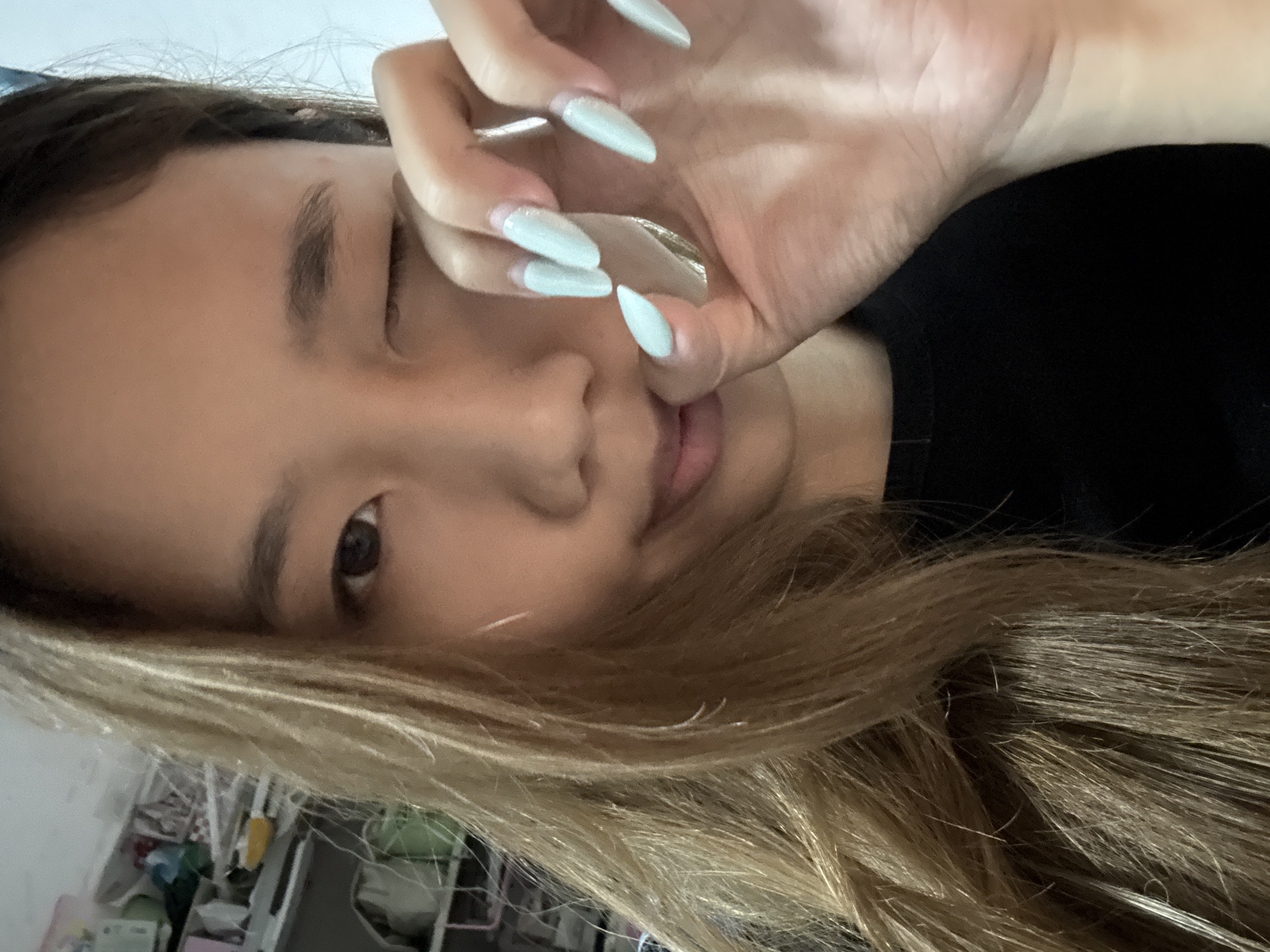 A young Asian woman with long, blonde hair is in a close-up shot, appearing to be indoors. She is winking with her left eye, while her right eye is open. Her right hand is raised to her face, with her fingers touching her lips. She has long, almond-shaped fingernails painted a light blue color. She is wearing a black top. The background appears to be a shelf with various items, suggesting a cluttered environment. The image is likely taken in Nan'an District, China.