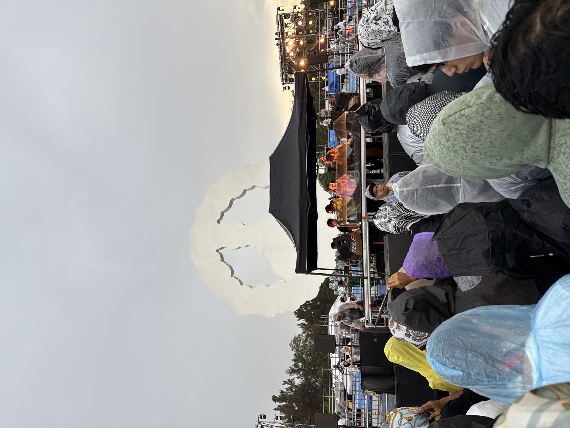 This image depicts an outdoor event in Mexico City, Mexico, likely a concert or festival, attended by a large crowd. The weather appears overcast and possibly rainy, as many attendees are wearing clear or colored plastic ponchos. The foreground is dominated by the backs and heads of the audience, showing a variety of hoods and hairstyles. In the mid-ground, there is a stage area covered by a black tent, with several people standing behind what appears to be DJ equipment. Behind the stage, a large, white, cloud-like sculpture is visible against the sky. Further back, there are lighting rigs and stage structures, suggesting a professional setup for a performance. The overall atmosphere suggests an event in progress despite the weather.