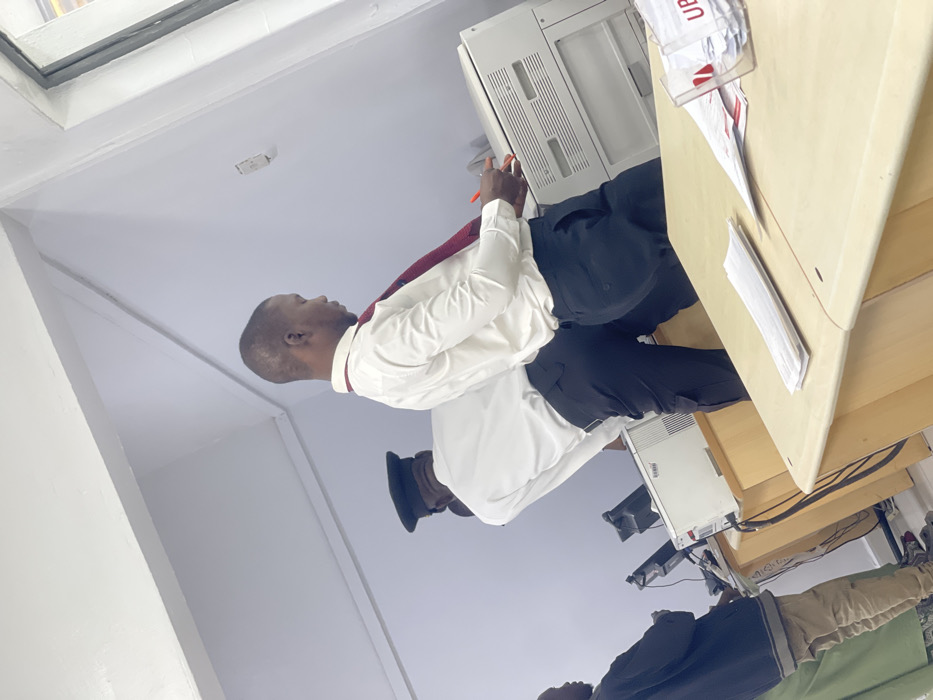 The image depicts an indoor office or administrative area in Zaria, Nigeria. Three individuals are present within a brightly lit space featuring white walls and a light-colored ceiling with visible support beams.
In the mid-ground, a male subject with short hair, wearing a white long-sleeve shirt, a dark red tie, and dark trousers, stands with his back partially to the camera, leaning towards a large, light-colored multifunction copier machine. He holds a red pen or marker in his right hand near the copier's control panel. Directly behind him, another male subject, also in a white long-sleeve shirt and dark trousers, wears a dark peaked cap. He is partially obscured and appears to be standing at a workstation with a computer monitor. Further to the left, a third individual, mostly visible from the waist down, wears a dark jacket, a light green garment, and light-colored trousers, standing at another workstation with a computer monitor.
The foreground features a light-colored desk or counter with stacks of white papers and a clear plastic holder containing additional papers, with the partial letters "UBE" visible on it. Multiple computer monitors and other office equipment are visible on desks throughout the scene. A white electrical outlet is mounted on the wall to the far right, adjacent to a visible window frame.