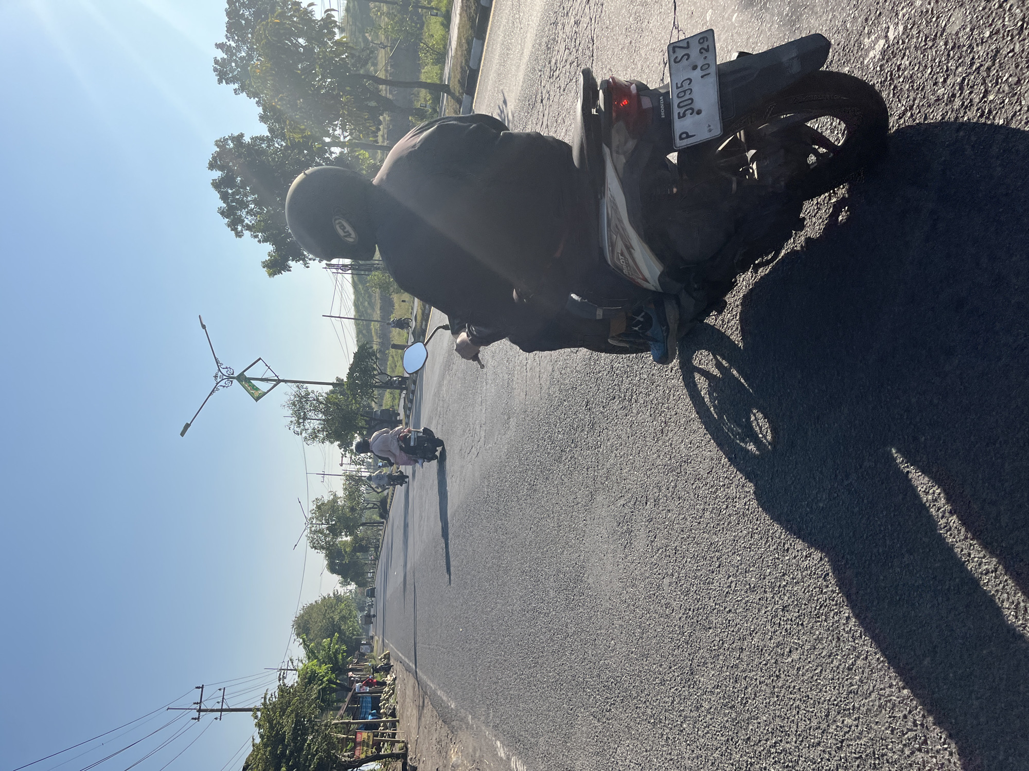 The image shows a rear view of a person riding a scooter on a paved road in Siring, Indonesia. The rider, wearing a black helmet with an "Arai" logo and a dark long-sleeved shirt, is positioned in the foreground, facing away from the viewer. The scooter is a white and beige Honda model with a license plate reading "P 5095 SZ 10.29". The road is asphalt, with some cracks and a white line marking the edge of the lane. Sunlight casts a long shadow of the scooter and rider across the road.
In the background, other scooters and riders are visible further down the road, suggesting a common mode of transportation. The environment includes trees lining the right side of the road, some with lush green foliage, and on the left, a roadside area with stalls or shops and more trees. Electrical poles and wires are present along the left side of the road, indicating an urban or semi-urban setting. The sky is a clear, bright blue. The overall impression is a sunny day on a busy road in Indonesia.