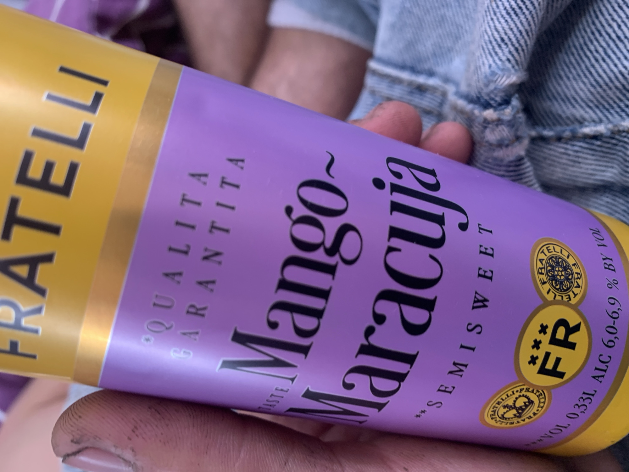 A person's hands are holding a tube of Fratelli Mango Maracuja drink. The tube is primarily purple with a yellow band at the top. The text "FRATELLI" is visible on the yellow band in black, block letters. The purple section of the tube has text in black and white. "QUALITA GARANTITA" appears in white, smaller text. The main text on the tube reads "Mango Maracuja" in large, stylized black lettering. Below this, "SEMI SWEET" is written in smaller black text. There are two circular emblems on the right side of the tube. The top one is yellow with a black, intricate design, and appears to say "FRATELLI" around its edge. The bottom emblem is also yellow and contains the letters "FR" in black, with three asterisk-like symbols above the "F". Further text along the bottom right of the tube reads "*VOL 0.33L ALC 6.0-6.9% BY VOL". The background is blurred and shows hints of denim fabric and a striped pattern, possibly indicating clothing or furniture. The lighting suggests it is daytime. The image is cropped closely to the tube and the hands holding it.