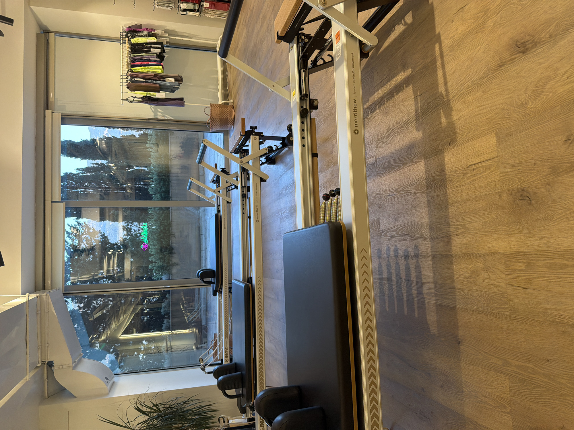The image shows the interior of a Pilates studio in Boreti, Montenegro. The studio features several Pilates reformers, primarily white with black padded surfaces. Two reformers are prominently displayed in the foreground, one partially obscured. The machines have wooden accents and metal components. In the background, a large window offers a view of trees and a building with a neon sign. A rack of athletic apparel is visible to the right of the window, along with a woven basket. The floor is made of wooden planks. The lighting suggests it is daytime, with natural light filtering in from the window and some artificial light from the ceiling. There are no people visible in the image.