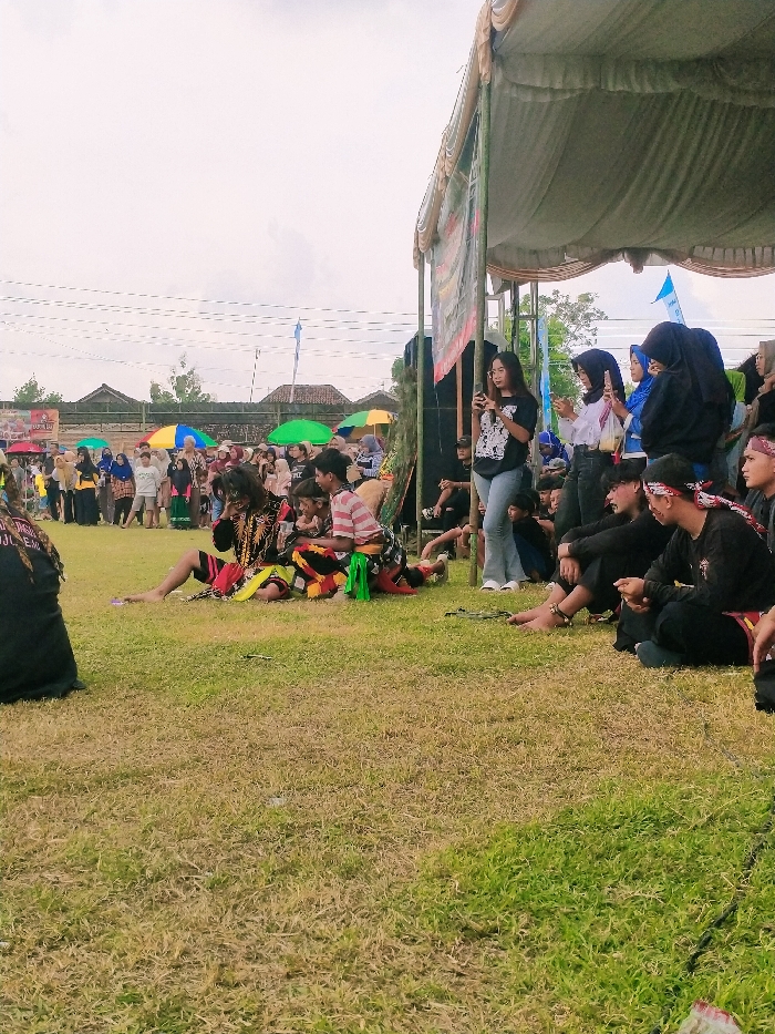 The image shows a gathering of people in an open-air space in Karangmalang, Indonesia (latitude: -7.47, longitude: 111.064). The scene appears to be a performance of traditional dance or theater. Several men, dressed in traditional costumes, are seated on the grass. The men are performing a dance or play, as indicated by their stylized gestures. A group of people are watching the performance, sitting on the grass or standing under a large white canopy tent. Several women are standing and watching, some holding smartphones or other devices. There are various colorful umbrellas in the background. The environment is characterized by grass, trees, and some buildings. The image is taken from a slightly elevated viewpoint, looking down on the scene.