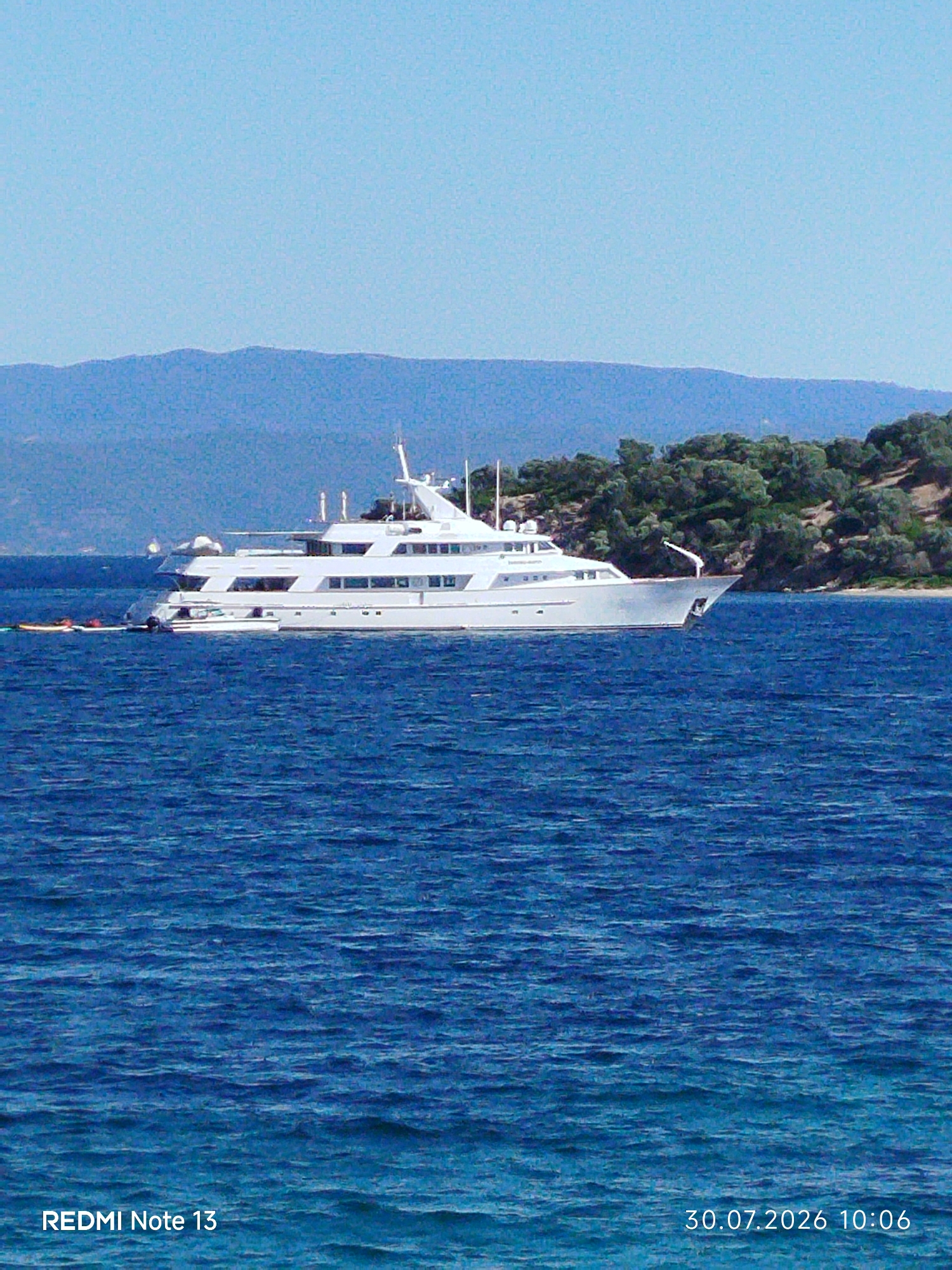 A wide, long shot captures a large, white luxury yacht at anchor in the clear blue waters off the coast of Sithonia Municipal Unit, Greece. The yacht is positioned in the mid-ground, with its sleek, multi-decked structure dominating the frame. A smaller tender boat is visible attached to the yacht's stern. In the background, a verdant, tree-covered hillside slopes down to the water's edge, with hazy, blue mountains forming the distant horizon. The sky above is a bright, clear blue. The image is dated "30.07.2026 10:06" and bears the watermark "REDMI Note 13".