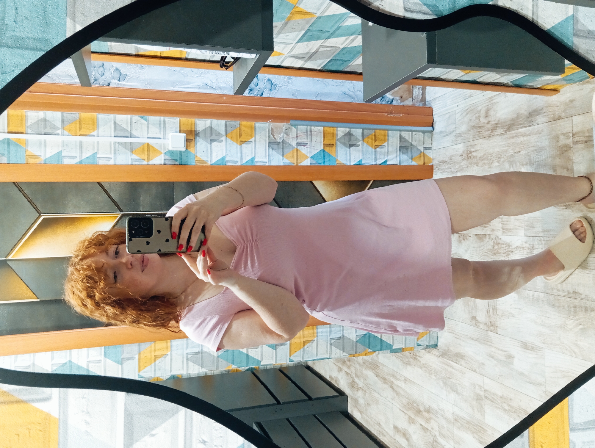 A red-haired woman in a pink dress takes a selfie in a mirror. She holds a phone with a black and beige dotted case and has red nail polish. She is wearing a gold bracelet on her left wrist and tan sandals. The background includes a wall with a pattern of geometric shapes in blues, yellows, and grays, wood trim, and a floor with wood-grain texture. A piece of black furniture is also visible. The location is Yurgamysh, Russia.