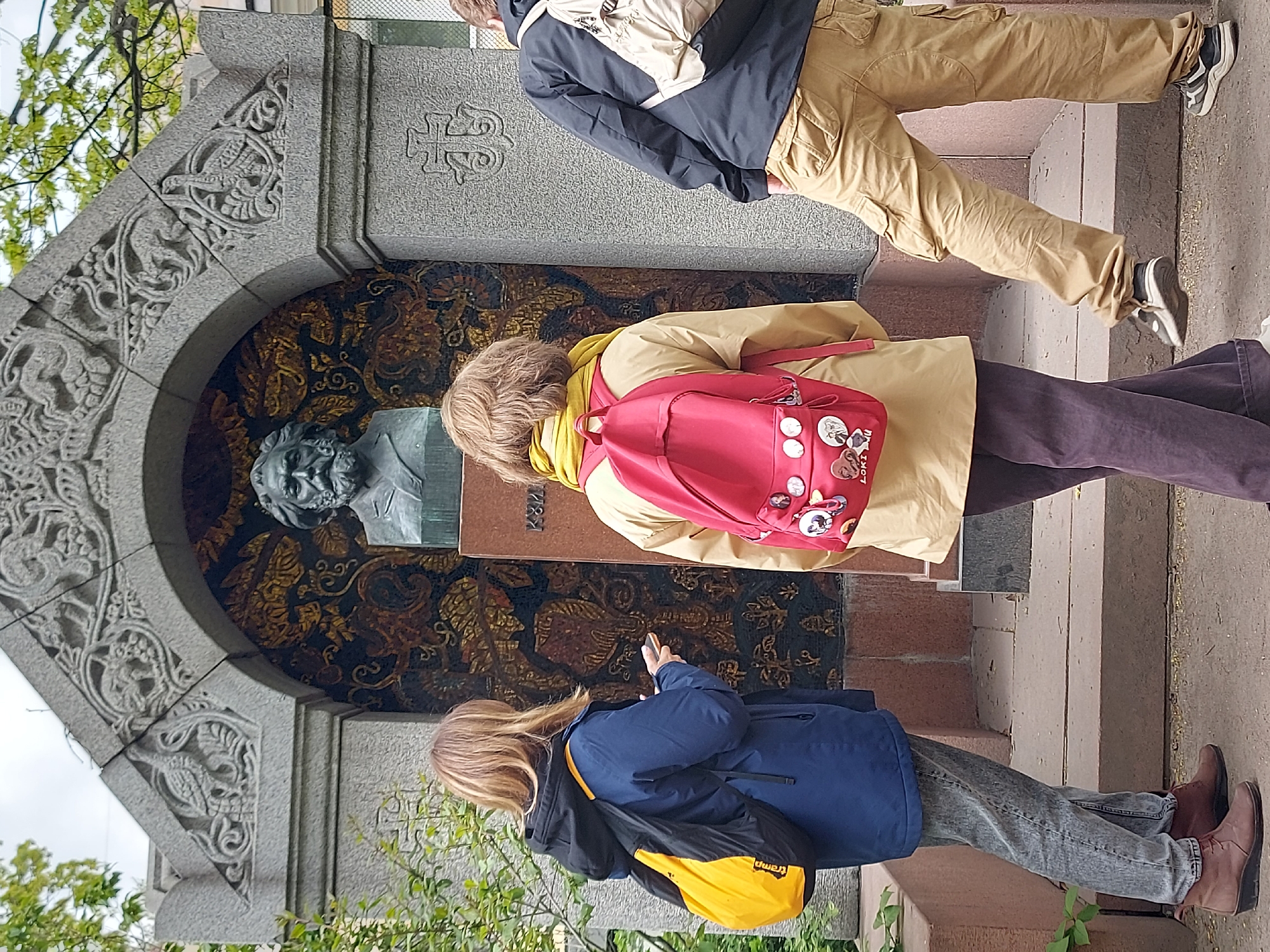 The image shows three individuals standing in front of a monument in what appears to be a cemetery or memorial park in Saint Petersburg, Russia. The monument is an arched structure with intricate carvings. Within the arch is a dark background with a mosaic pattern featuring gold, brown, and black, and a bronze bust of a person.
The three individuals are all standing on a set of steps leading up to the monument.
* The person on the left is facing the monument. She is wearing a blue jacket, grey jeans, and brown boots and has a yellow and black bag.
* The person in the middle is wearing a tan jacket, brown pants, and a red backpack with multiple pins.
* The person on the right has tan pants and a black jacket.
The surroundings suggest a well-maintained outdoor space, likely a significant cultural or historical site.