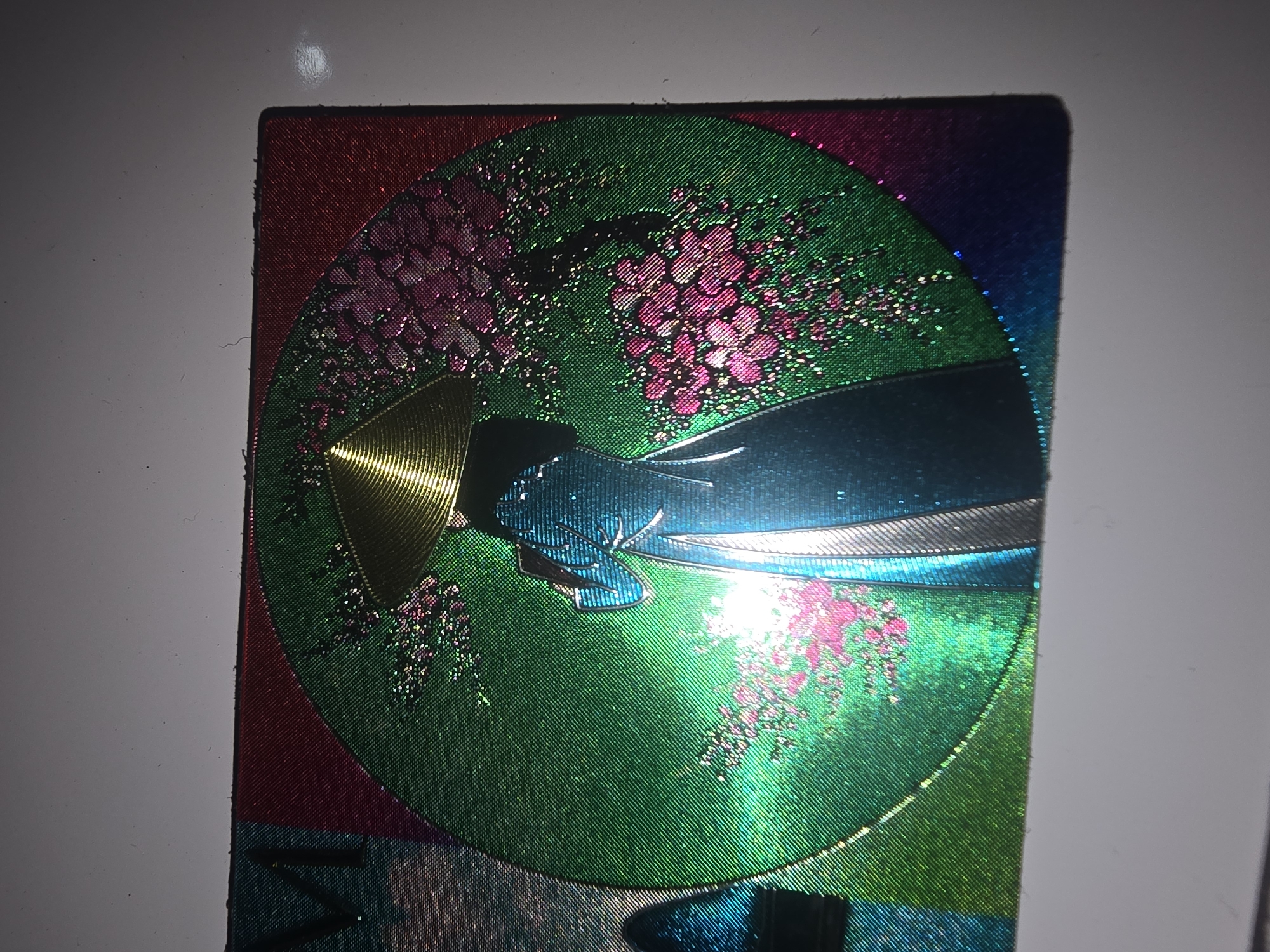 The image displays a close-up view of a holographic sticker. The sticker features an illustration of a woman in traditional Vietnamese attire, wearing an ao dai and a conical hat, standing under a blooming cherry blossom tree. The background is a vibrant green circle, with accents of pink and red at the edges, suggesting a sunset or a festive backdrop. The holographic effect causes the colors to shimmer and shift with the light. The sticker appears to be adhered to a flat, light-colored surface, likely a table or a wall. There are no people visible in the immediate vicinity of the sticker. The image is not related to Ulan-Ude, Russia.