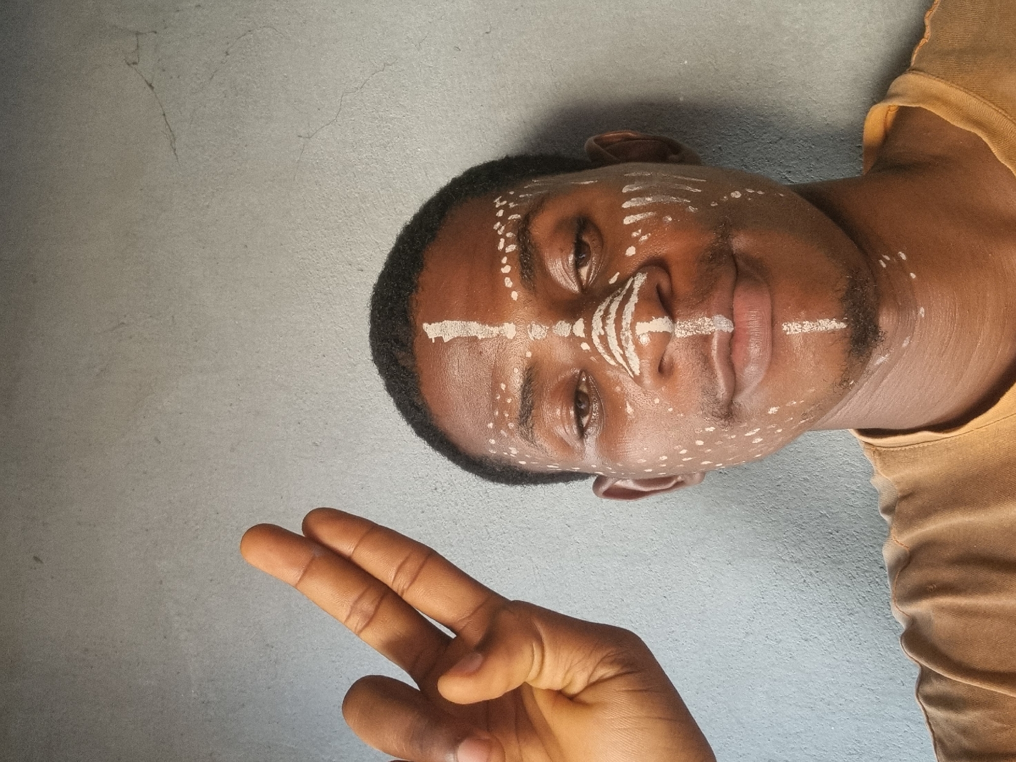 An upward-facing portrait features a person with dark skin and short dark hair, adorned with intricate white face paint, against a plain light gray wall background. The person's head is tilted to the right, and their gaze is directed towards the viewer with a subtle, gentle expression.
The face paint consists of several prominent white lines: one across the forehead, one spanning the bridge of the nose and extending onto the cheeks, another above the upper lip, and a short line on the chin. Numerous small white dots are scattered across the forehead, temples, cheeks, and below the eyes, complementing the linear patterns.
In the lower-left foreground, a hand, presumably the subject's, is visible, making a "peace" sign with the index and middle fingers extended. The person is wearing a mustard yellow or light brown t-shirt, visible on their right shoulder and neck area. The background wall shows some minor textural imperfections and faint cracks. The lighting is soft and even, highlighting the details of the face paint. There is no visible text.