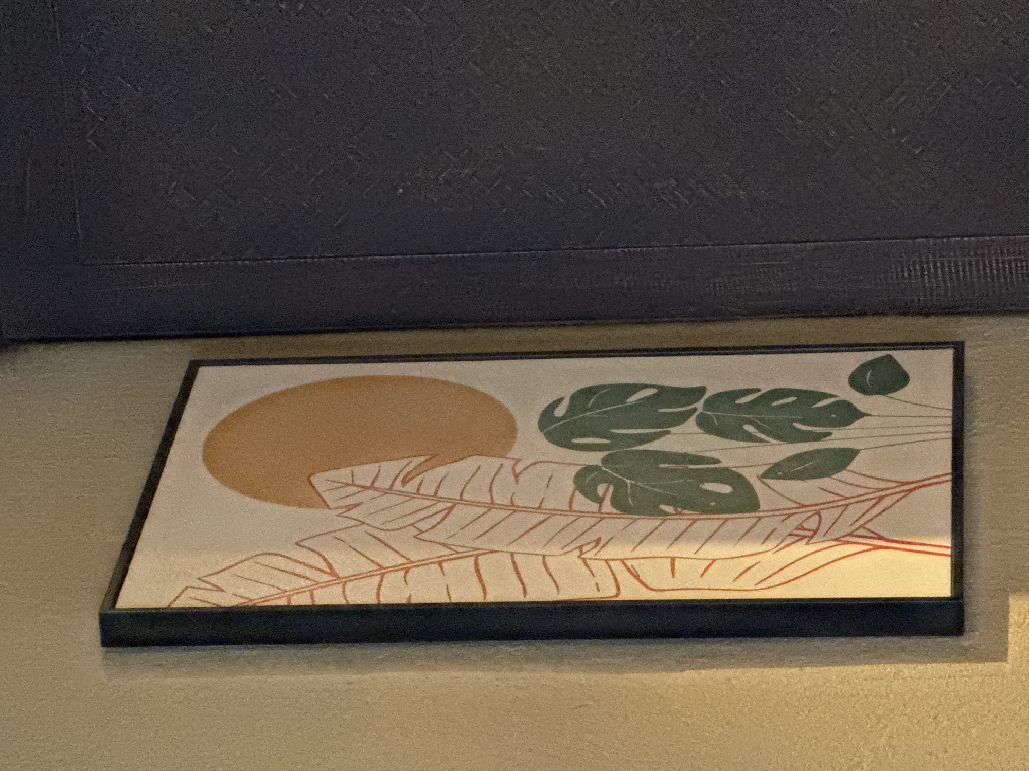 An framed abstract artwork featuring stylized green leaves and orange plant stalks against a pale background with a large orange circle representing the sun is leaning against a light tan textured wall. To the right of the artwork, a dark blue textured wall is visible, forming a corner with the tan wall. The scene is dimly lit, with a warm light casting shadows and highlighting the textures of the walls and the artwork. The image is taken from a slightly upward angle. There are no people or animals present. The location is not discernible from the image.