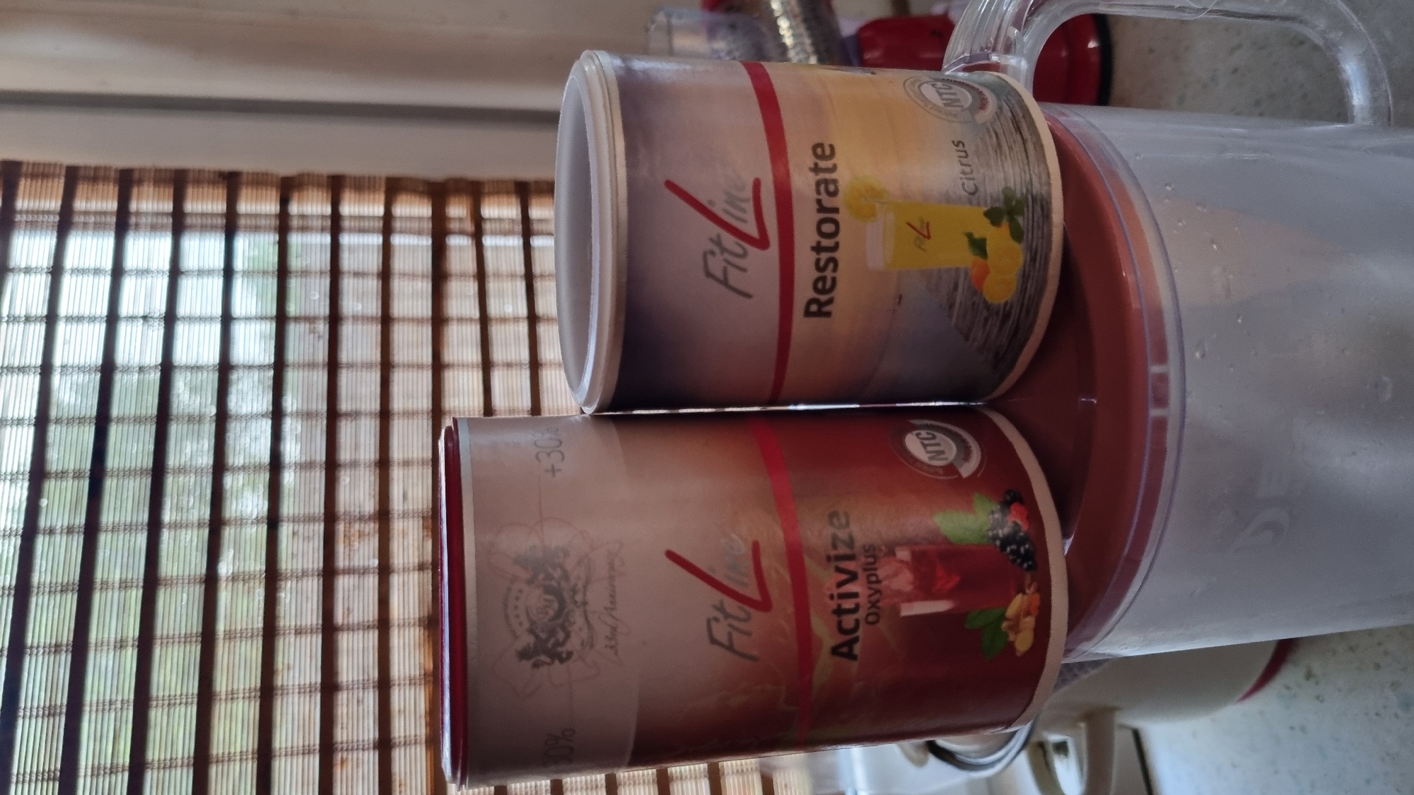 The image shows two cylindrical containers of "FitLine" products, likely dietary supplements, placed on top of a transparent pitcher. The container on the left is labeled "Activize Oxyplus" and features an image of a red beverage with berries and mint. The container on the right is labeled "Restorate" and depicts a yellow drink with lemon and mint. Both containers have the "FitLine" logo prominently displayed. The background consists of horizontal blinds, suggesting an indoor setting. The scene is set in Солнечная Поляна-3 Снт, Russia.