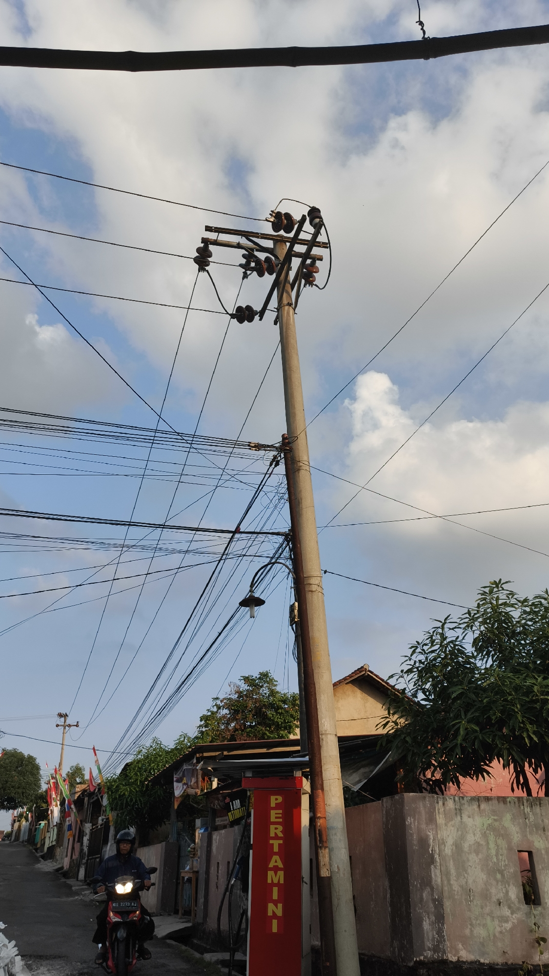 This image depicts a street scene in Bandar Lampung, Indonesia, featuring a tall, weathered utility pole laden with numerous electrical wires. The pole, centrally located and angled slightly to the right, supports multiple sets of insulators. A streetlamp is attached to the pole lower down.
To the left of the pole, a person wearing a dark blue jacket and helmet rides a red motorcycle, identified by the license plate "BE 3273 AJ". They are traveling down a paved street lined with houses and small structures. A red gasoline pump, labeled "PERTAMINI" in yellow vertical lettering, stands prominently to the right of the street.
Behind the gasoline pump and to the right of the utility pole, a section of a concrete wall is visible, adjacent to a building with a pinkish-red exterior and a tiled roof. Lush green trees, including a large one with dense foliage, grow around the structures. The sky above is a clear blue, dotted with scattered white clouds, suggesting it is daytime. Several flags are visible on poles along the left side of the street, adding to the local Indonesian atmosphere. The overall scene captures a typical roadside view in a residential area of Bandar Lampung.