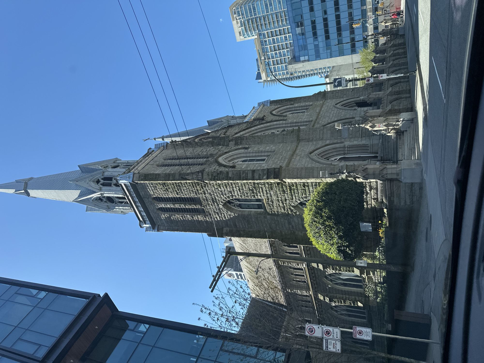 The image shows Christ Church Cathedral in Vancouver, Canada, viewed from a street-level perspective. The architectural style of the cathedral is Gothic Revival, constructed from grey stone. The cathedral features a tall steeple, pointed arches, and multiple windows with intricate tracery.
The church is juxtaposed with modern architecture, including a tall skyscraper in the background. Street signs and a utility pole are present in the foreground. A trimmed green bush is situated near the building. The sky is clear and blue. The perspective suggests the photograph was taken from a moving vehicle.
