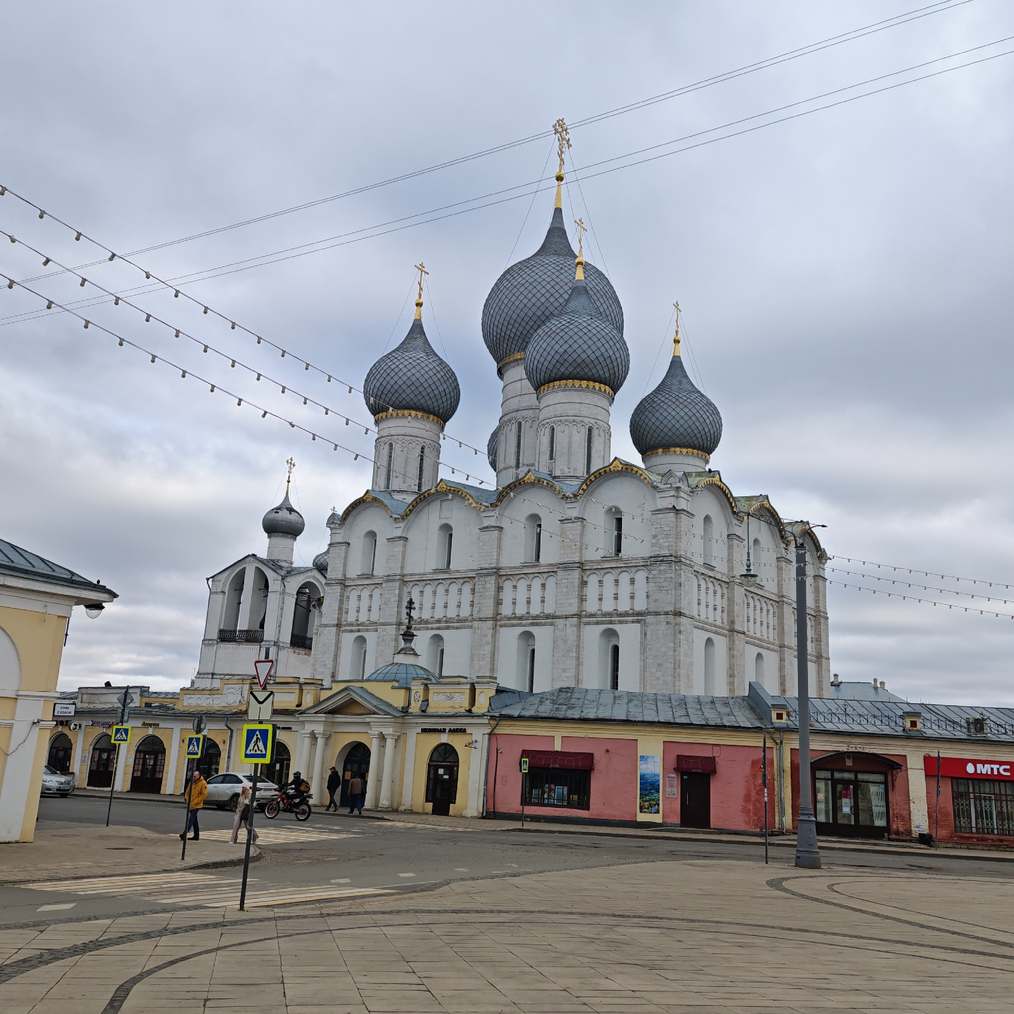The image captures a cityscape in Rostov, Russia, featuring the Rostov Kremlin. The main focus is the Assumption Cathedral, a large white stone building with five prominent, grey, onion-shaped domes topped with golden crosses. The cathedral is surrounded by lower structures, including buildings with pink, yellow, and white facades, housing shops and commercial establishments, one of which is branded "MTC."
In the foreground, there is a pedestrian crossing with several pedestrian crossing signs. A man in a yellow jacket is walking across the crosswalk, and a motorcycle is also present. The ground is paved with light grey paving stones with dark grey curved lines forming patterns. The sky is overcast with grey clouds. Overhead, there are strings of lights, adding to the urban ambiance.