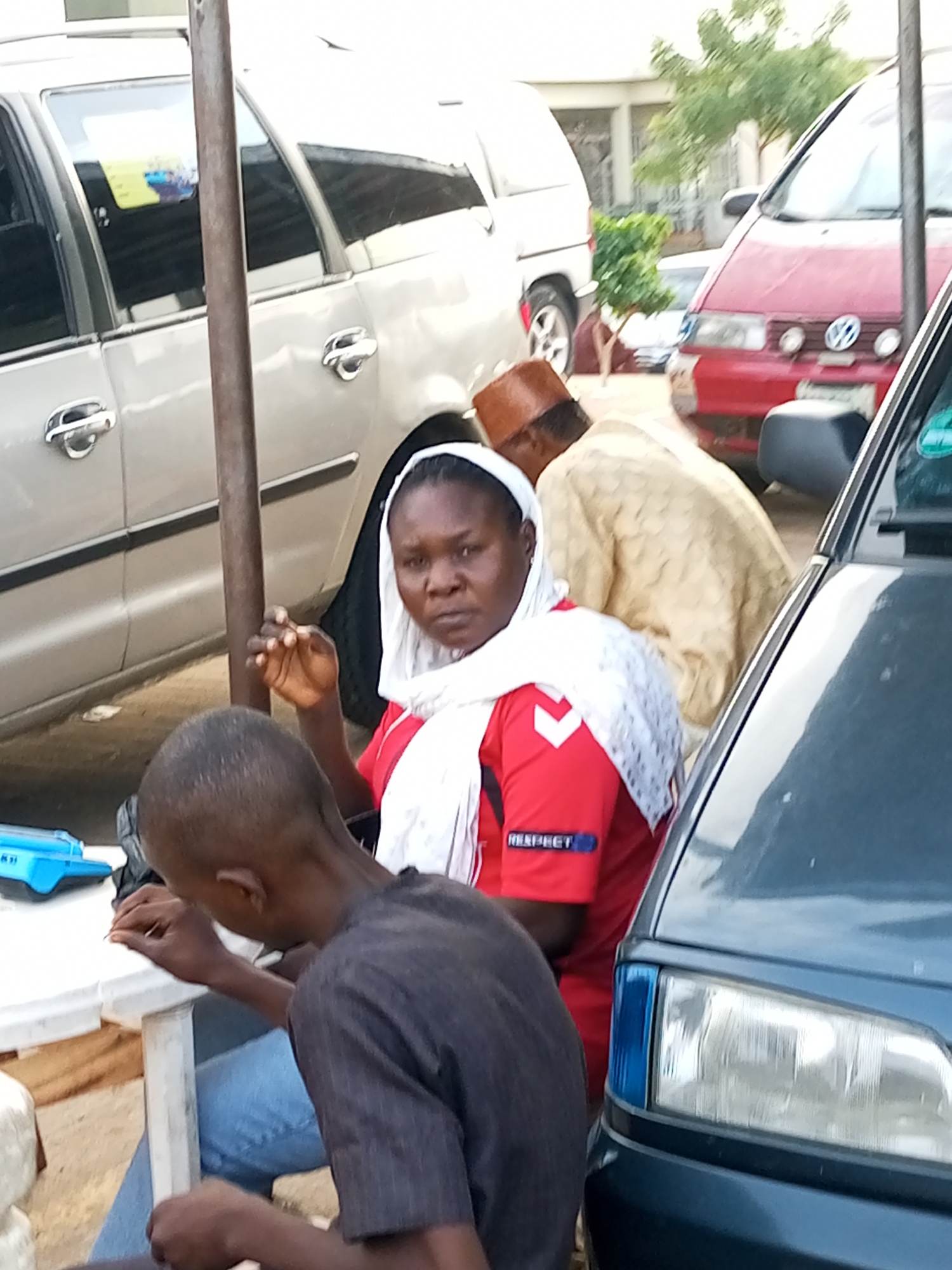 In Gombe, Nigeria, a group of people are gathered near vehicles. A woman in a red shirt and white headscarf is seated at a white table, looking forward with a neutral expression. Next to her, a young person in a dark grey t-shirt and blue jeans is leaning over the table, appearing to be engaged in a task. Behind them, a man in a light brown patterned garment and a brown hat is visible. The scene is framed by several vehicles, including a light-colored SUV on the left, a red Volkswagen on the right, and a dark-colored vehicle in the foreground. The environment appears to be an outdoor area, possibly a roadside or market, with a pole in the center of the frame and trees and buildings in the background. The lighting suggests it is daytime.