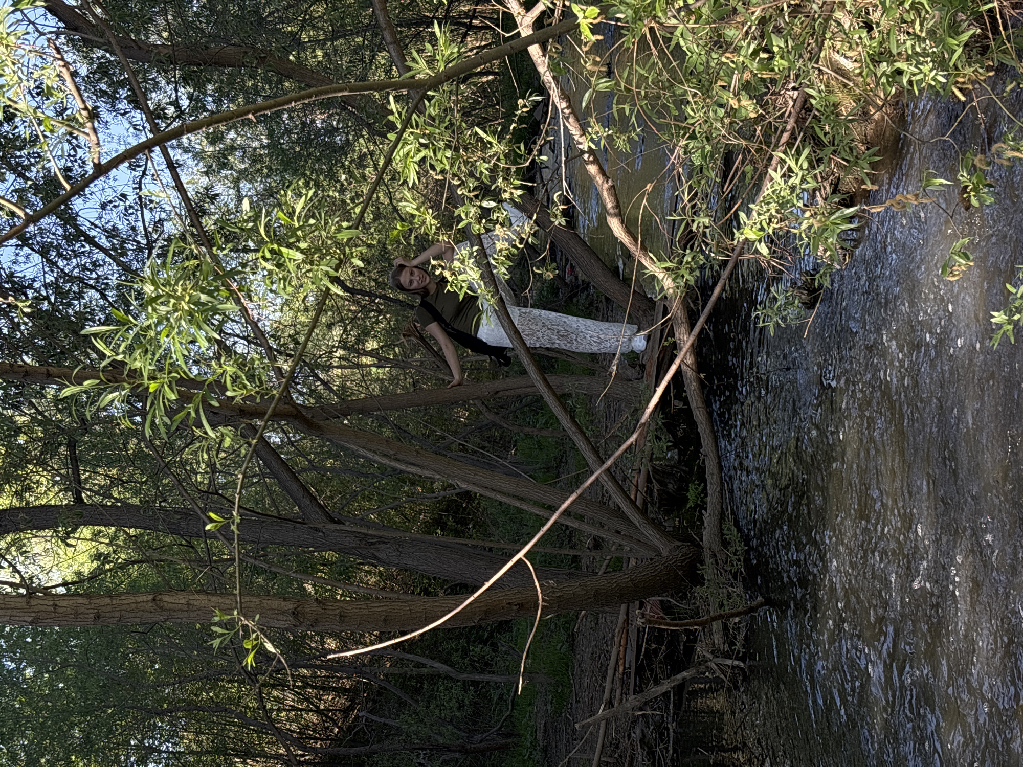 A young woman stands on a fallen tree branch over a body of water, possibly a river or stream. She is wearing an olive-green shirt, white lace pants, and black shoes. A black bag is slung over her shoulder. Her left hand is on her head, and she is looking at the camera. The water is dark with sunlight reflecting on its surface. The scene is surrounded by trees and foliage. The location may be in or near the city of Perm, Russia.