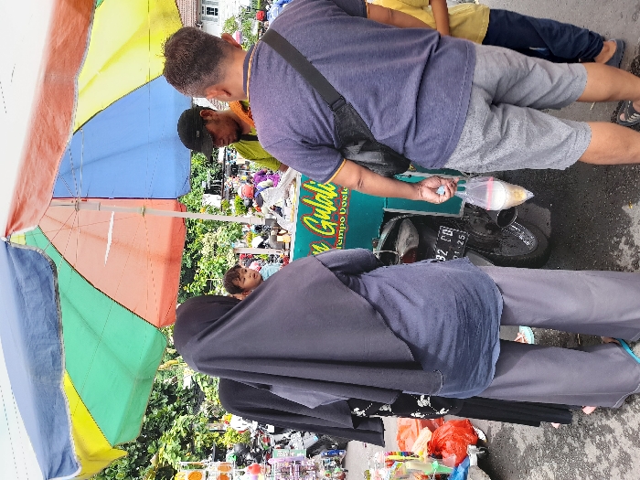 The image shows a street food vendor in Kecamatan Sidoarjo, Indonesia (latitude: -7.441, longitude: 112.705). A man wearing a blue shirt and a black satchel is holding a plastic bag with a beverage inside, standing next to a food cart with an umbrella. The cart has a sign that reads “Gulabi Tempo Doeloe”. A woman in a black hijab and a blue shirt is standing next to him. Another woman wearing a dark grey shirt and black hijab is standing behind the woman in blue. A small child is standing in front of the woman in blue. There are several motorcycles parked in the background. The scene is set on a busy street with parked motorcycles and other street food vendors visible in the background. The street is lined with trees and buildings.
