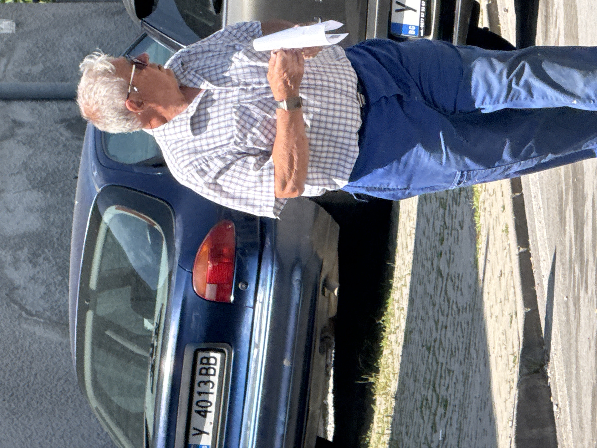 An elderly man stands beside a blue car, holding a piece of paper. The car's license plate reads "Y 4013 BB." The scene appears to be on a city street with cobblestone and concrete surfaces. The city and region are not identifiable.