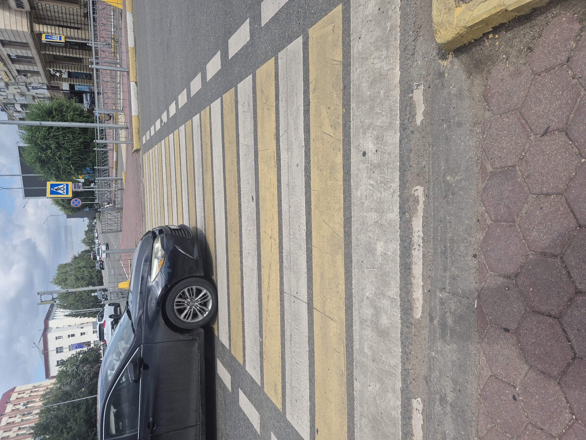 A black sedan is stopped at a pedestrian crossing on a city street. The crossing has alternating yellow and white stripes. The car's front passenger-side wheel is positioned on the first yellow stripe. The road is asphalt and appears to be in good condition. Beyond the crossing, a sidewalk with red hexagonal paving stones is visible, bordered by a raised curb. In the background, there are buildings, trees, and other vehicles on the road. The sky is overcast. The scene appears to be in Kokshetau, Kazakhstan, as suggested by the presence of pedestrian crossing signs with blue backgrounds and white figures, which are common in the region.