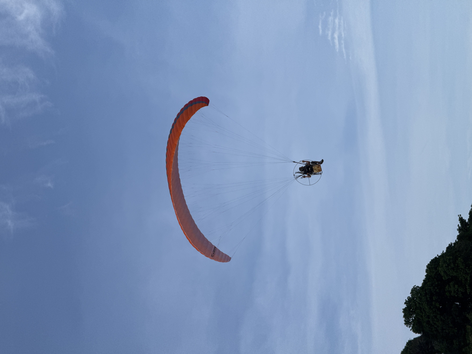 An airborne paramotor is centrally positioned against a light to medium blue sky featuring scattered wispy and diffused cloud formations. The paramotor utilizes an inflated orange canopy with a darker band along its trailing edge; the brand name "OZONE" is discernible on the underside of the canopy's right wingtip. A single pilot, dressed in dark attire, occupies a seated position within a harness and frame assembly, which includes a visible circular propeller guard and a rear-mounted engine unit. Numerous suspension lines connect the canopy to the pilot's rig. In the lower-left quadrant of the image, dense dark green tree foliage is visible. The scene is situated in Rawai, Thailand.