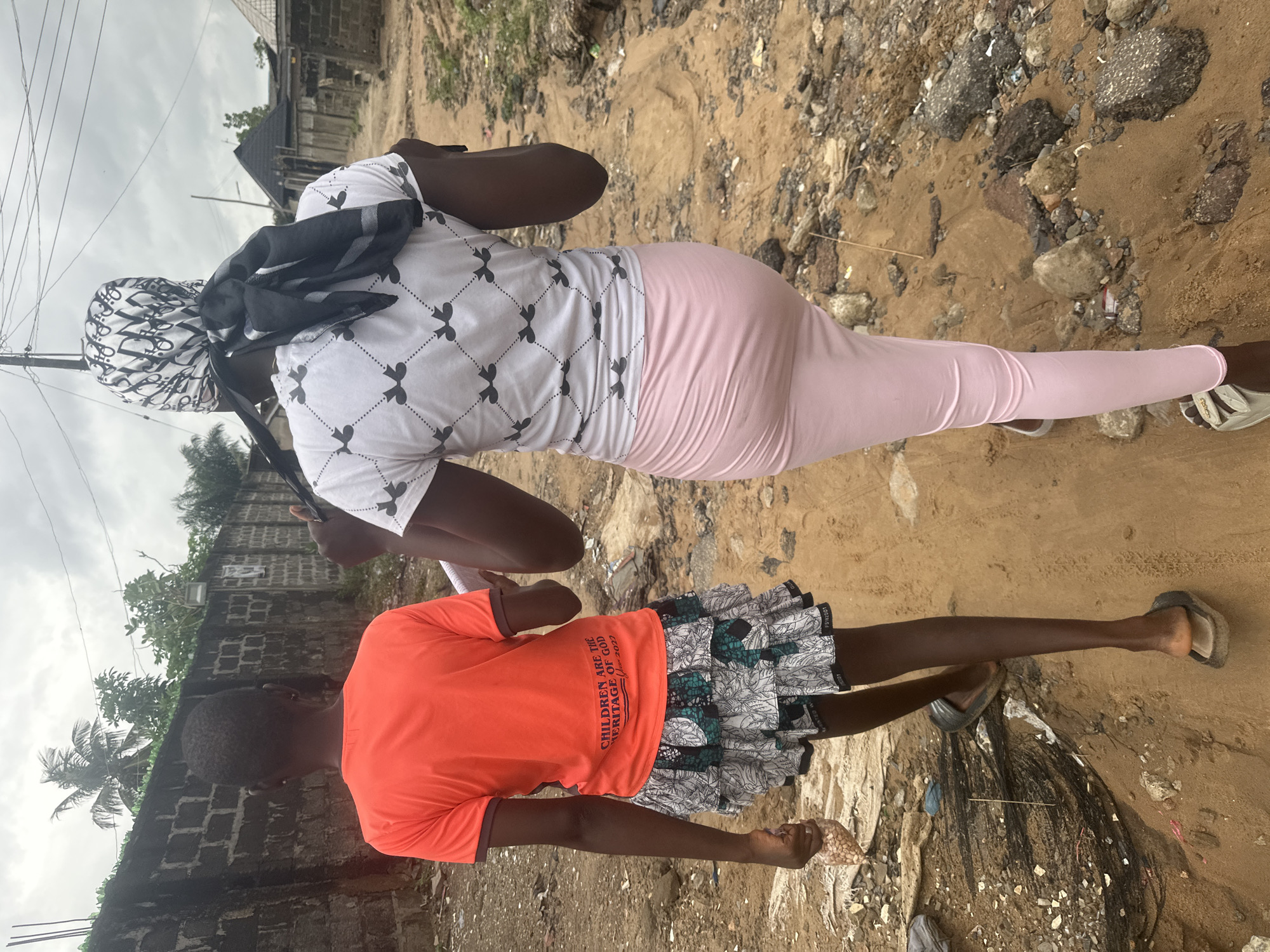 In the city of Ajara, Nigeria, two individuals, viewed from behind, are walking away from the camera on a dirt path. The person on the left is wearing an orange t-shirt with text that reads "CHILDREN ARE THE HERITAGE OF GOD Year 2022," and a patterned skirt. They are carrying a small plastic bag of what appears to be nuts in their left hand. The person on the right is wearing a white t-shirt with a repeating pattern of black bird-like figures and a light pink skirt or leggings. A black and white patterned scarf is tied around their head, with the ends hanging down. The environment is characterized by a rough, sandy terrain with scattered debris, including rocks and what looks like broken asphalt. A brick wall runs along the left side of the path, with trees and vegetation visible above it. A utility pole is present in the background. The sky is overcast.