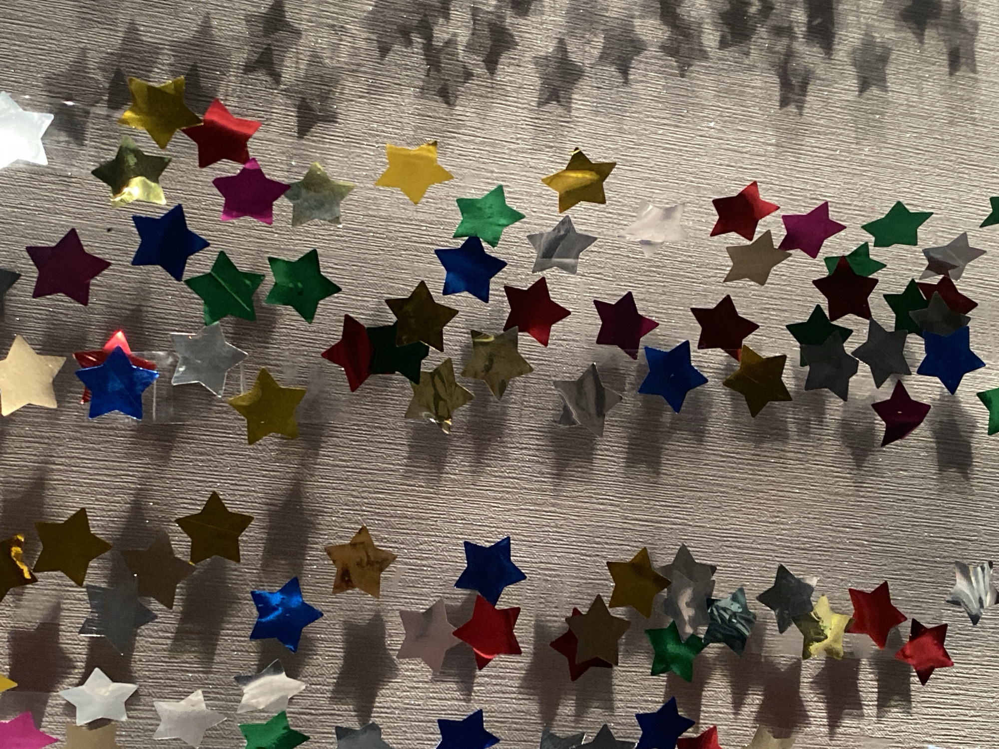 The image is a close-up, high-angle shot of a textured, light-colored surface with numerous small, multi-colored star-shaped confetti pieces scattered across it. The confetti appears to be made of a shiny, metallic material and is depicted at various angles and depths, creating a sense of randomness and visual interest. The lighting casts subtle shadows from the confetti onto the textured surface. The colors of the confetti include red, blue, green, yellow, silver, white, and a deep magenta or purple. There are no people or identifiable landmarks present in the image. The location is Unknown, Unknown.