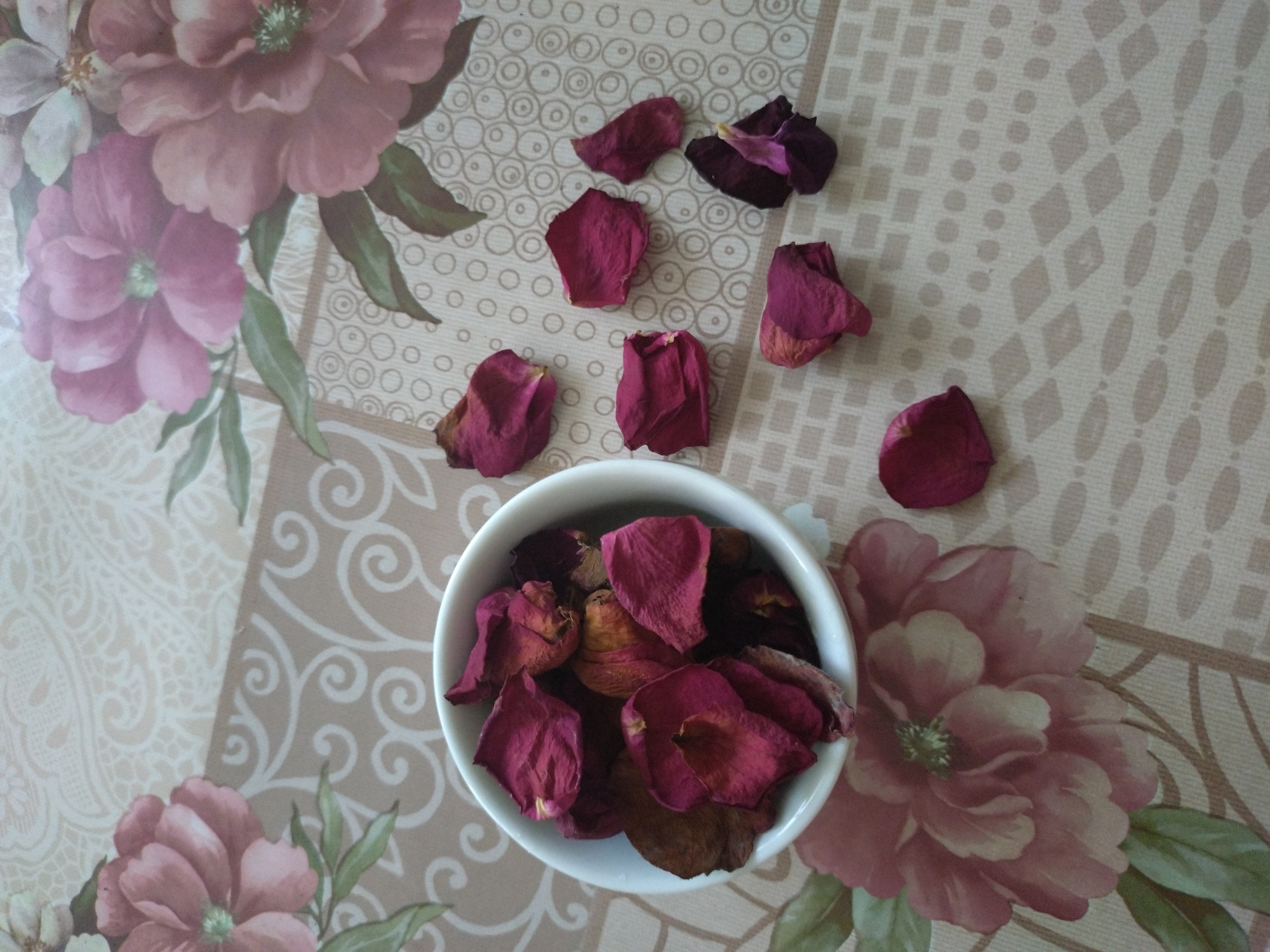 The image provides a top-down view of dried rose petals arranged on a patterned tablecloth, captured in Stara Zagora, Bulgaria. A small, white ceramic bowl, positioned slightly below the center, is filled with a collection of deep red and burgundy rose petals, some exhibiting warmer orange and brown hues indicative of their dried state. Several individual petals of similar color are scattered loosely on the tablecloth around the bowl.
The tablecloth itself features a detailed design. On the left side, there are large, soft pink and mauve floral patterns with green leaves. The right side and background areas display various geometric motifs in light beige and brown tones, including patterns of concentric circles, ovals, and squares. The lighting is soft and natural, suggesting daytime. No specific activity or event is depicted beyond the presentation of the dried petals. There is no visible text in the image.