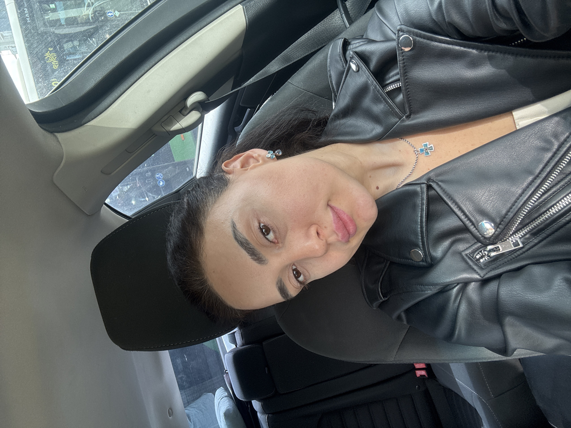 A woman is seated in a car, wearing a black leather jacket and a seatbelt. She has dark hair and is wearing jewelry: earrings and a necklace with a cross pendant. The car's interior is visible, including the headrest and other seats. The location appears to be Tbilisi, Georgia based on text visible outside the car window.