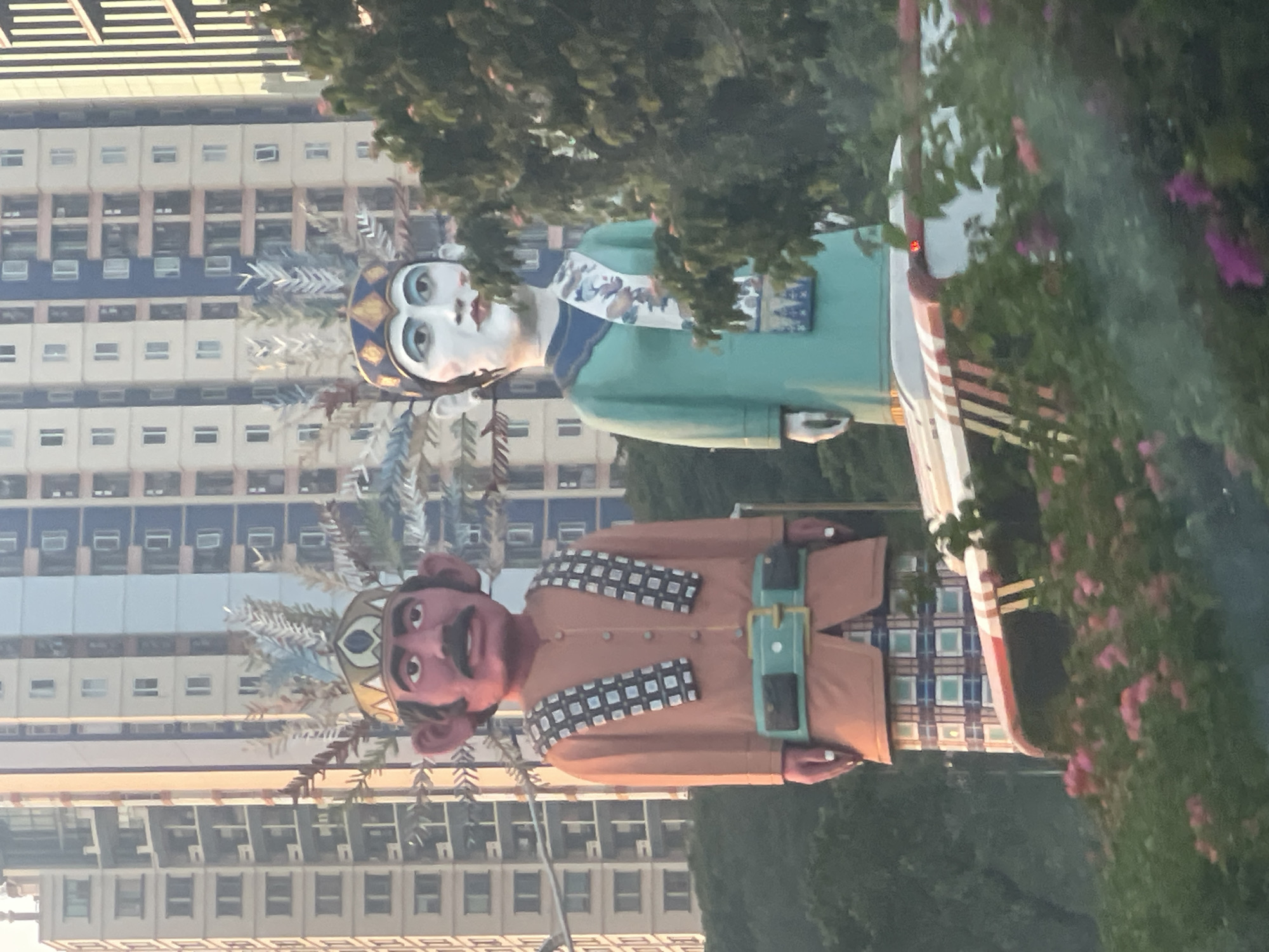 Two large, effigy-like figures are positioned in the foreground, in front of a backdrop of tall, modern buildings. The figure on the left has red skin, a mustache, and large ears, wearing an orange garment with a patterned scarf and a wide belt. The figure on the right has pale skin and is wearing a teal garment with a patterned sash. Both figures appear to be wearing ornate crowns. Behind them, several multi-story buildings with a grid-like pattern of windows dominate the scene. In the lower portion of the image, green foliage and pink flowers are visible, partially obscuring a vehicle with a red and white striped canopy. The lighting suggests it is daytime, possibly late afternoon given the warm tones on the buildings. This scene is located in Central Jakarta, Indonesia, where such figures, known as Ondel-ondel, are often displayed.