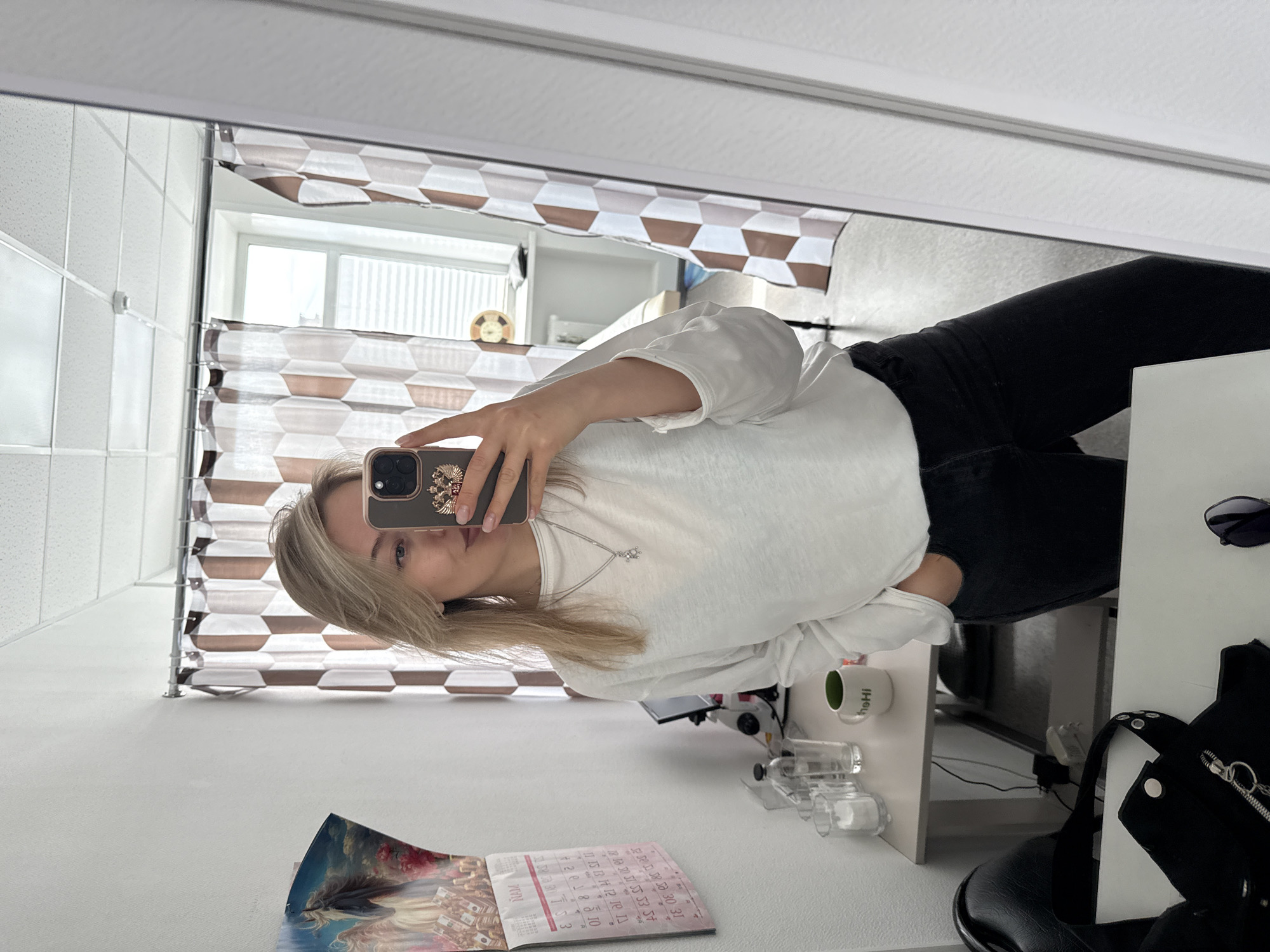A fair-skinned woman with blonde hair takes a selfie in a mirror. The mirror reflects part of a room with a window and decorative curtains. The woman wears a white long-sleeved shirt and black pants. In the foreground, a table holds a calendar, a mug, and a black jacket. Sunglasses are placed on the table. The photo does not provide enough information to determine a specific location in Krasnoyarsk, Russia.