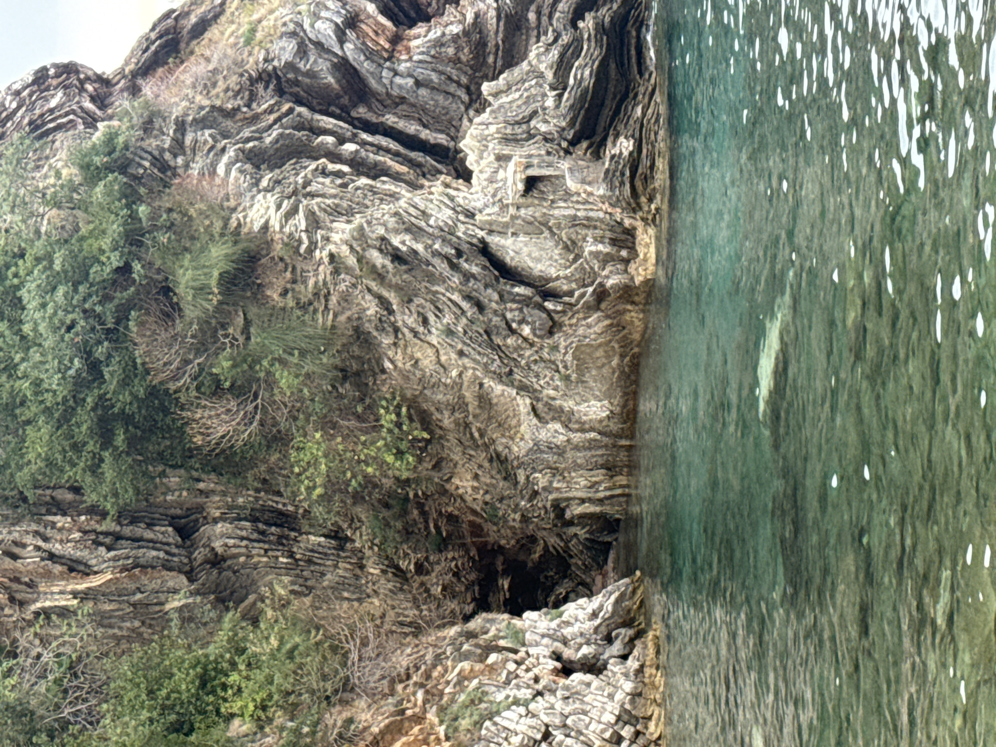 A rocky cliff face with a cave entrance and lush green vegetation rising above a clear, turquoise body of water. The water has a visible rock formation submerged beneath its surface. The rock face displays layered striations and appears weathered. The image captures a section of the coastline near Rafailovići, Montenegro.