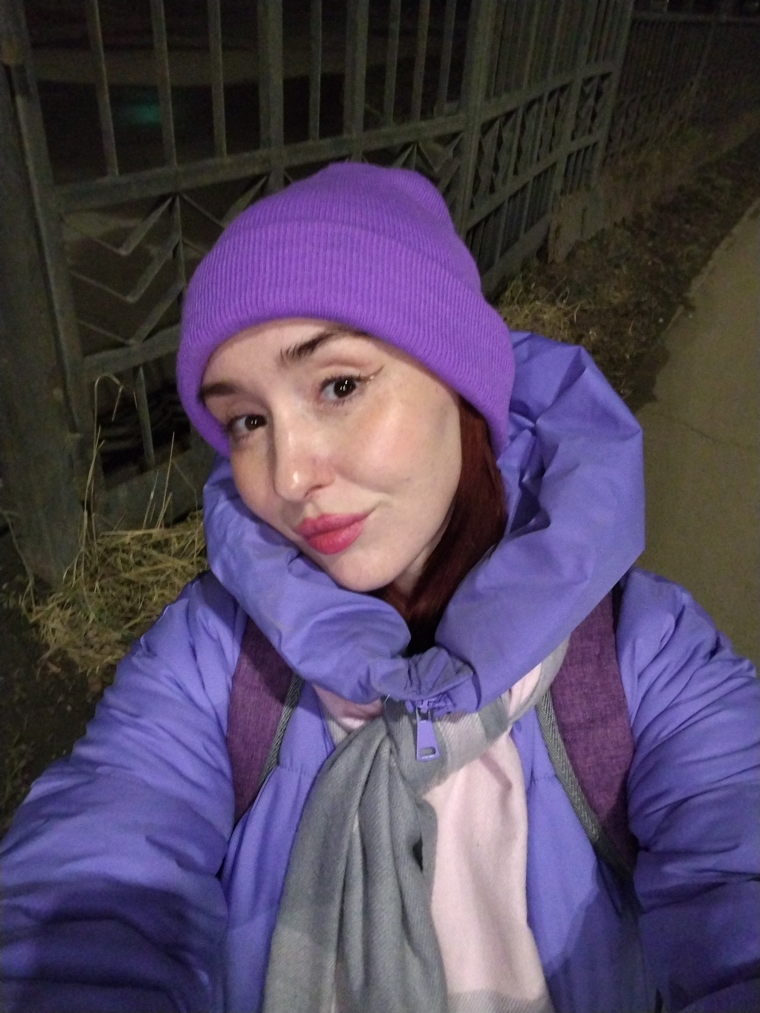 A person, presumably female with fair skin, red hair, and wearing a vibrant purple beanie and matching puffer jacket, is captured in a selfie. The individual has noticeable makeup, including eyeliner and pink lipstick. A grey scarf with pink accents is visible beneath the jacket. The background suggests an outdoor setting, possibly at night or under dim lighting. A metal fence runs horizontally behind the person, with dry grass and a sidewalk visible in sections of the background. The location is specified as Izhevsk, Russia.