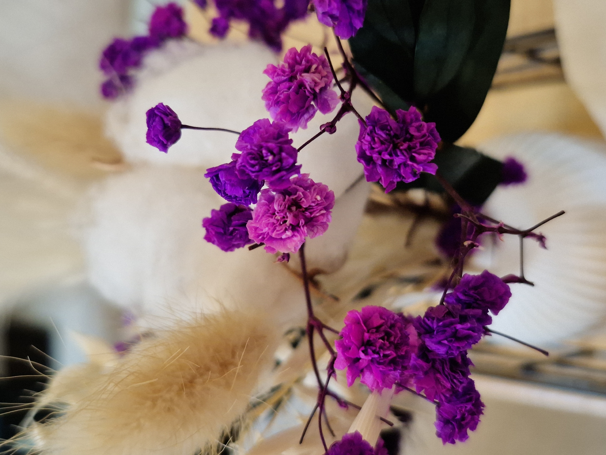 A close-up, eye-level, vertical, indoor shot features a bouquet of dried flowers. The focus is on the vibrant purple and fuchsia flowers, which are small and clustered on thin, dark stems. Behind them, softer, muted colors of dried pampas grass and white, fluffy elements create a textured background. The bouquet is arranged in a white, ridged vase, parts of which are visible at the bottom of the frame. The lighting is soft, highlighting the delicate textures of the flowers. The scene appears to be in an indoor setting, possibly a home or a studio, but no specific city or location is discernible.