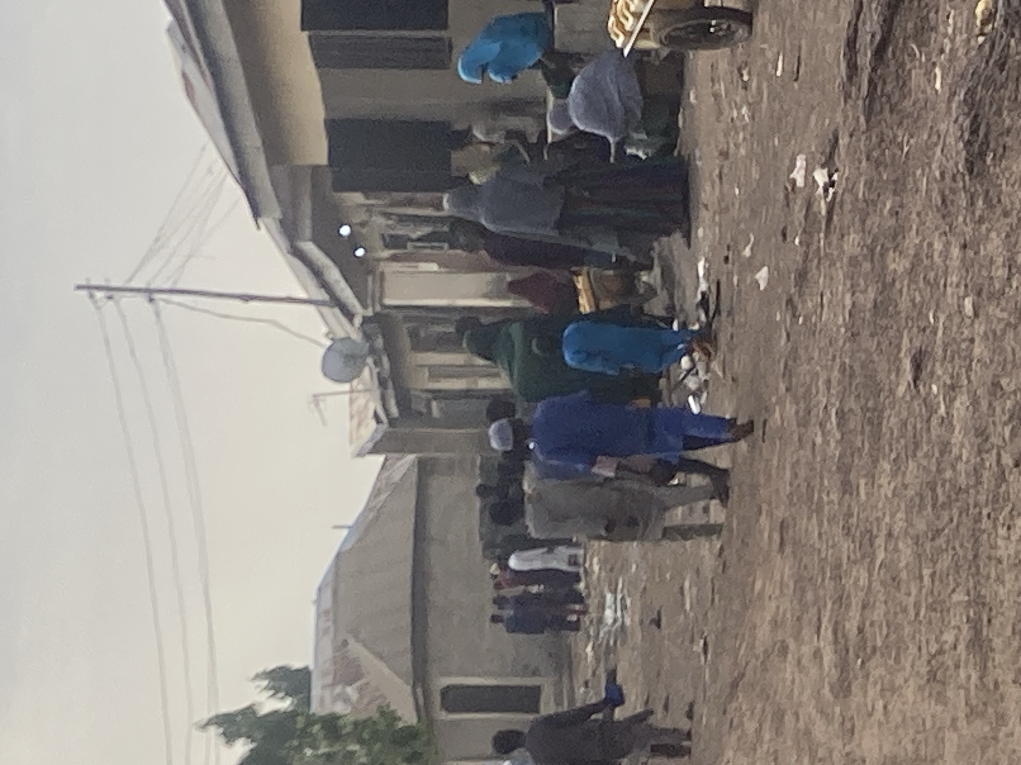 A street in Minna, Nigeria is depicted in this photograph. A group of people are walking along the street in both directions. Some individuals are wearing traditional clothing, including blue robes and caps. The street surface is littered with debris. Buildings line the street, with visible electrical wires above. A satellite dish is mounted on one of the buildings. The sky appears overcast.