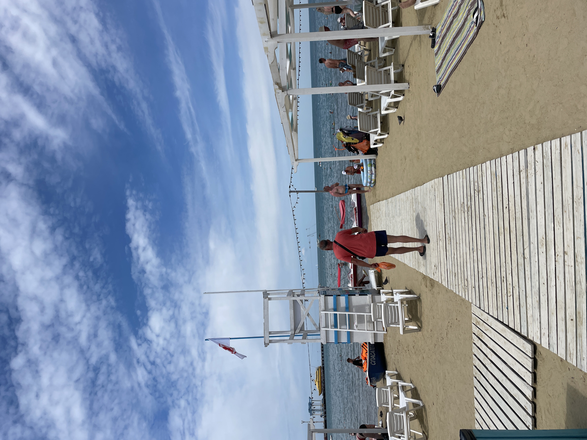A bright, sunny day at a beach in Anapa, Russia. A wooden walkway leads across the sand towards the calm sea. A man in a red shirt and blue shorts walks away from the camera, carrying an orange bag. Several beach umbrellas and lounge chairs are set up on the sand. In the background, people are swimming and playing in the water. A yellow ferry is visible in the distance. A lifeguard tower stands near the water's edge, with a flag flying at the top. The sky is a vibrant blue, with scattered white clouds.