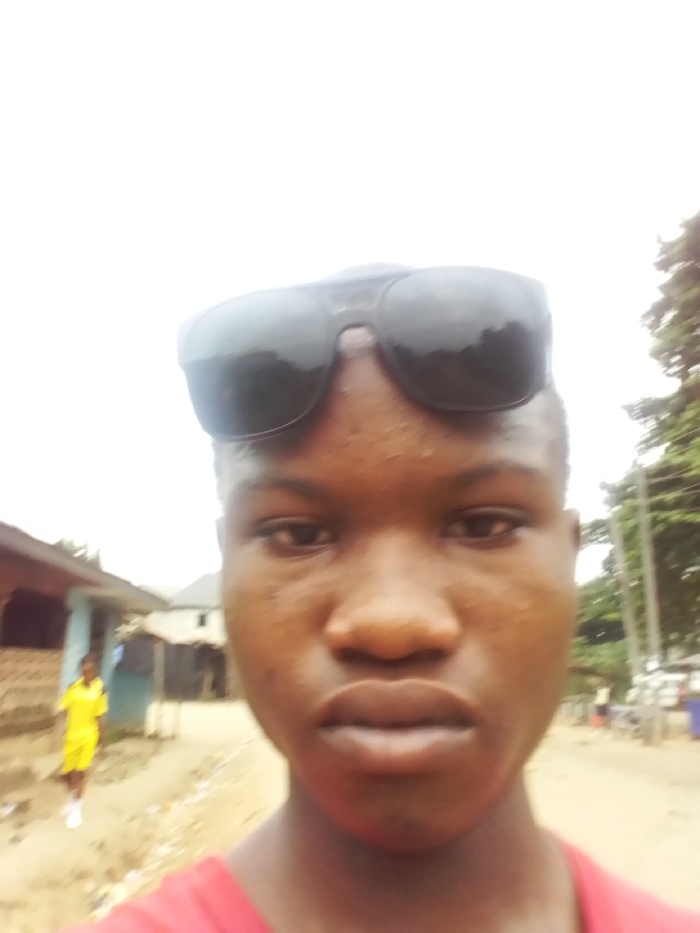 A close-up, frontal image captures a young male subject with brown skin, short dark hair, and dark sunglasses perched on his forehead. He wears a red garment, partially visible at the bottom of the frame. The background, significantly out of focus, indicates an outdoor, semi-urban environment in Owerri, Nigeria. To the subject's left, a blurred unpaved road or path extends past several modest buildings, including one with a light blue wall and another exhibiting a reddish-brown textured facade. A second individual, appearing to be a younger person, is visible in the mid-ground on the left, walking away from the camera, dressed in a bright yellow outfit and white shoes. To the subject's right, blurred green tree foliage and faint vertical structures resembling utility poles are present. The lighting is diffused, consistent with an overcast daytime sky. No discernible actions or interactions beyond the primary subject posing are evident.