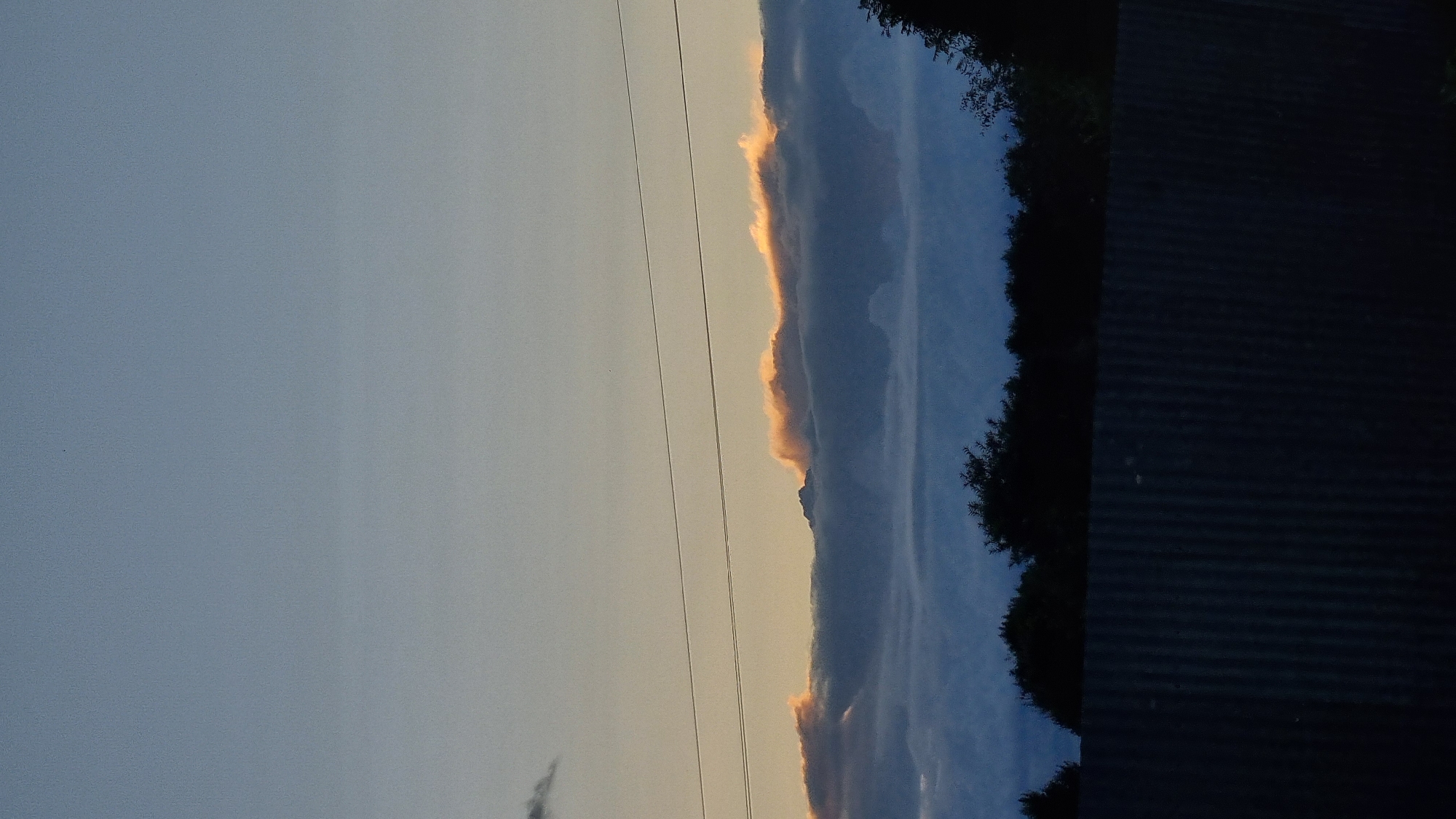 The image depicts a twilight sky over Vysokoe, Russia. A corrugated metal fence runs across the bottom foreground, casting the lower part of the scene in shadow. Above the fence, a dark silhouette of trees stretches across the horizon. The sky is transitioning from day to night, with a gradient of light blue and pale yellow, suggesting the sun is near or below the horizon. A long, horizontal band of clouds is visible, with the upper edge catching a warm, orange-pink glow, likely from the setting sun. Two thin, dark power lines are strung horizontally across the middle section of the sky.
