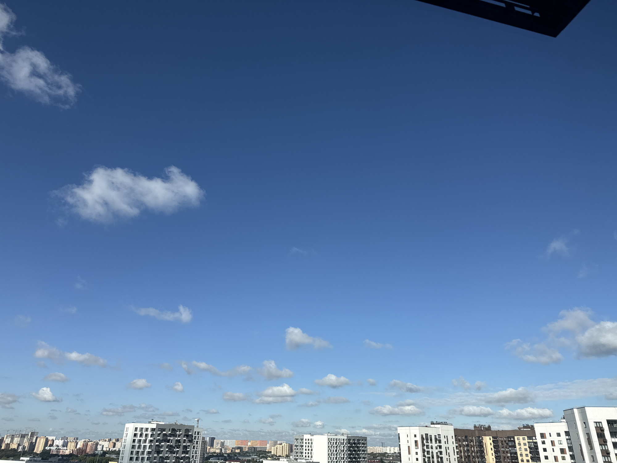 A wide shot captures a bright, clear blue sky with scattered white clouds above a cityscape in Lyubertsy, Russia. The horizon is dominated by a dense collection of modern residential buildings with varying architectural styles and colors, featuring numerous windows. A portion of a dark, angular structure, possibly a rooftop element or an overhang, is visible in the upper right corner of the frame. The buildings are densely packed, suggesting an urban or suburban environment. The overall scene depicts a daytime view with good visibility.