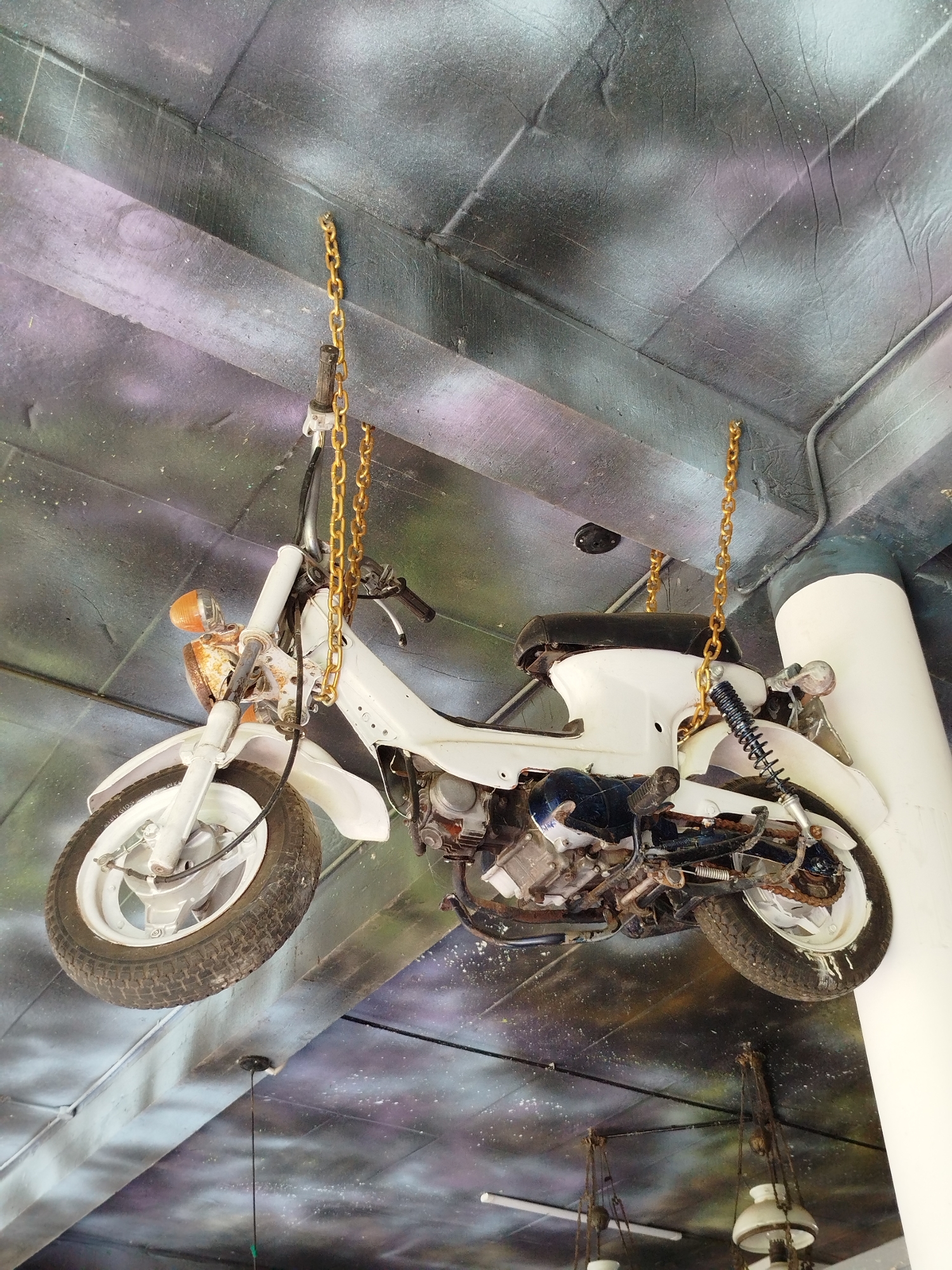 A white motorcycle hangs from the ceiling of a structure in Păng Tiêng 1, Vietnam. The motorcycle is suspended by two gold-colored chains attached to the handlebars and rear of the seat. The motorcycle has visible wear and tear, including rust and discoloration. The ceiling has a textured surface and a color scheme of gray, white, and purple. A white cylindrical column is visible in the background. Other hanging lights are present in the background.