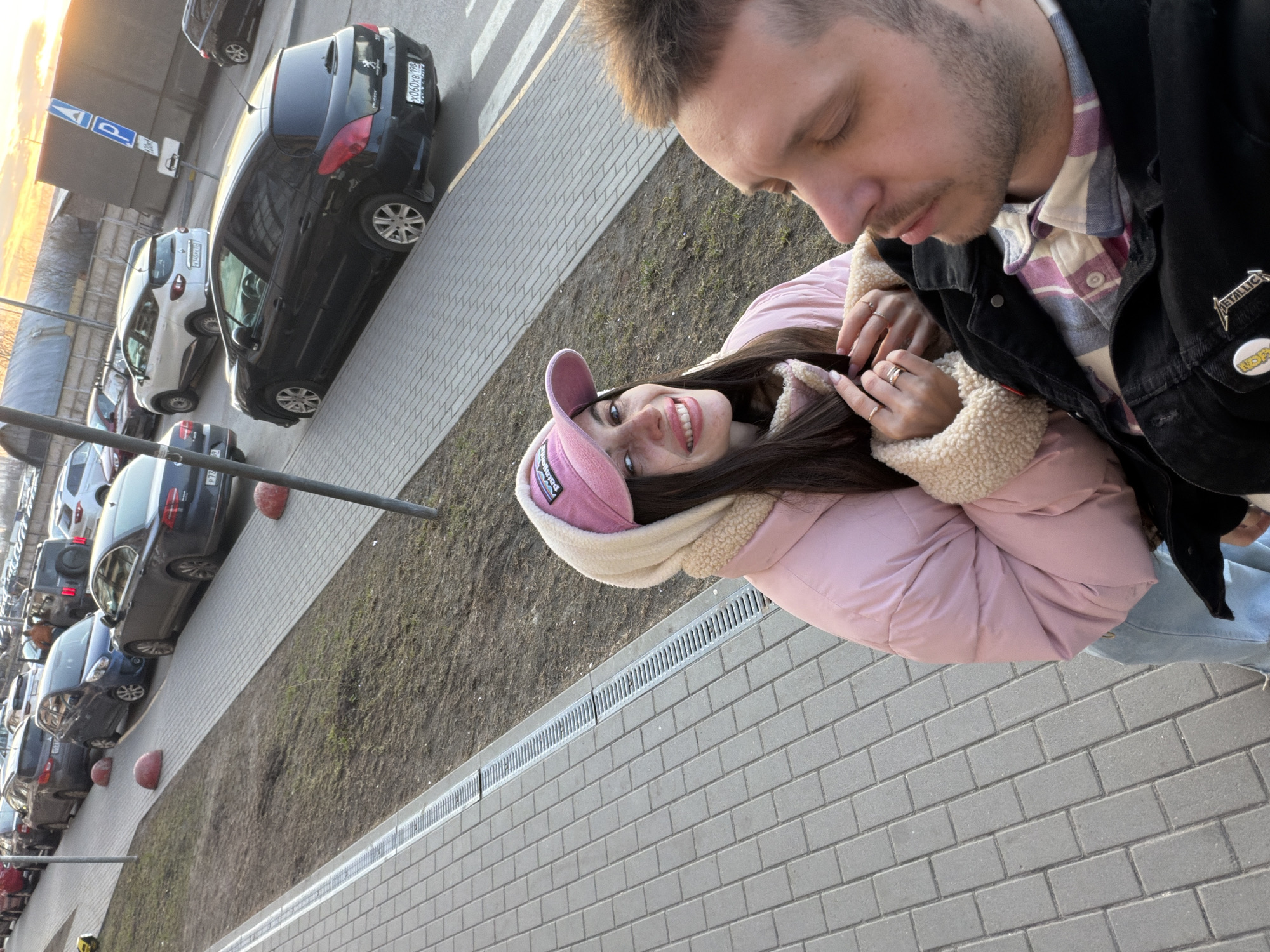 The image shows a man and a woman in an outdoor setting, likely Saint Petersburg, Russia, based on the license plate of the dark vehicle in the background. The woman, smiling at the camera, is wearing a pink jacket and a pink cap. The man, whose head is partially visible, is wearing a black jacket over a plaid shirt. They are standing on a brick-paved walkway. A drainage ditch runs along the walkway. A parking lot with multiple vehicles is visible in the background. The lighting suggests it could be early morning or late afternoon.