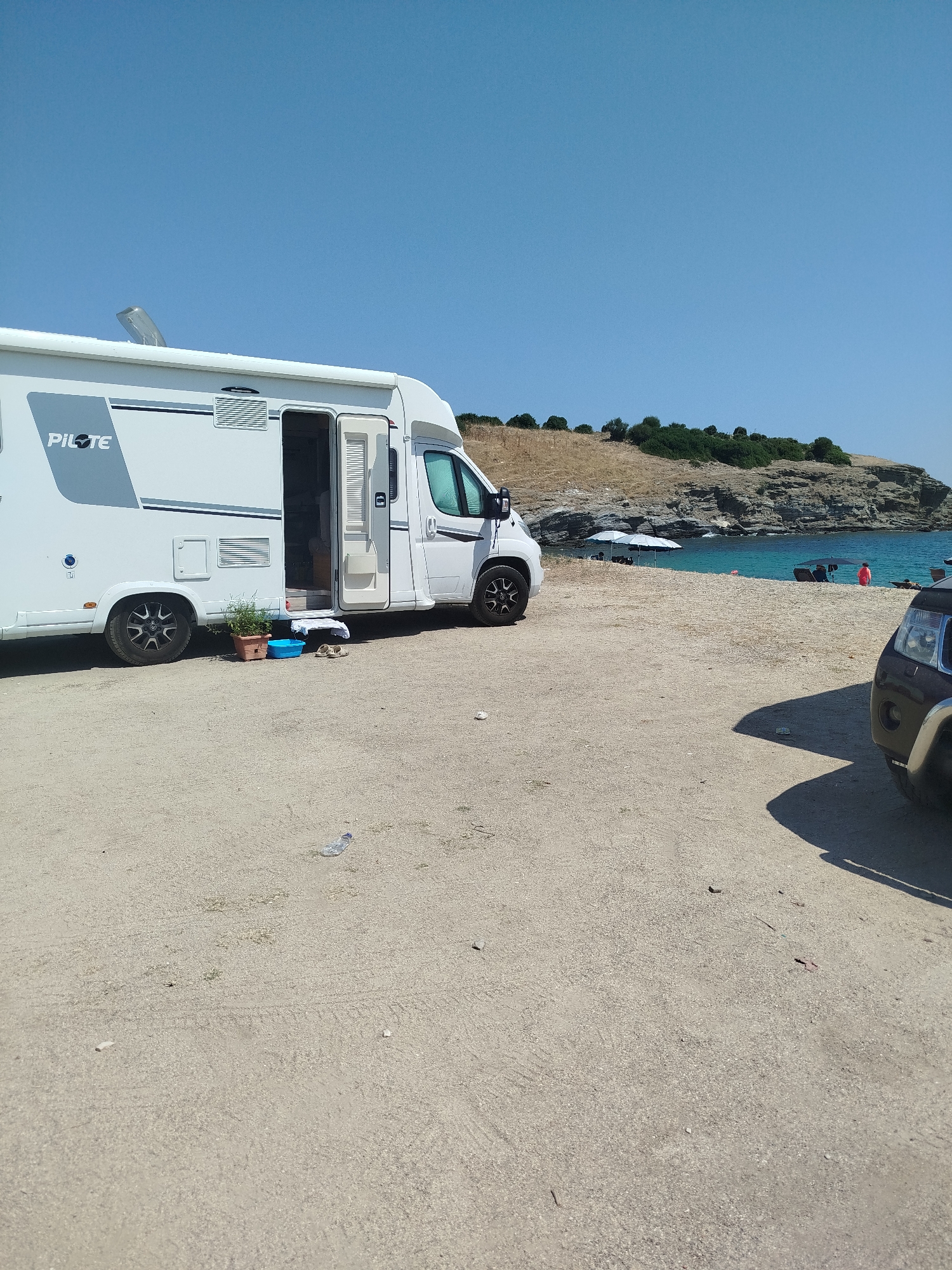 This image depicts a campsite on a sandy area near the coast of Sithonia Municipal Unit, Greece. A white camper van is parked on the left, with its door open, revealing steps leading inside. Next to the camper van are a potted plant and a blue basin. A dark-colored SUV is partially visible on the right side of the image, casting a shadow.
In the background, a clear blue sea meets a rocky shoreline with sparse vegetation. Several beach umbrellas and a few people are visible on the beach. The sky is a bright, cloudless blue, indicating a sunny day. The overall scene suggests a tranquil setting for camping and beach activities.