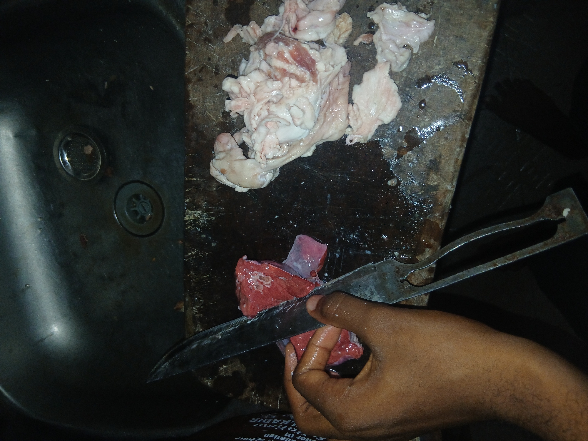 In Ikeja, Nigeria, a close-up shot captures a butcher's hands preparing raw meat in a dimly lit kitchen. The scene unfolds on a worn wooden cutting board, adjacent to a stainless steel sink. A person's left hand, with visible hair on the forearm, grips a large, sharp knife, poised to slice into a piece of red meat. The meat appears wet and glistens under the lighting. To the right of the red meat, a pile of white, fatty tissue and some red meat scraps are visible on the cutting board. The sink area, in the background, shows a drain and some water. The overall impression is one of a functional, albeit somewhat rustic, food preparation environment.
