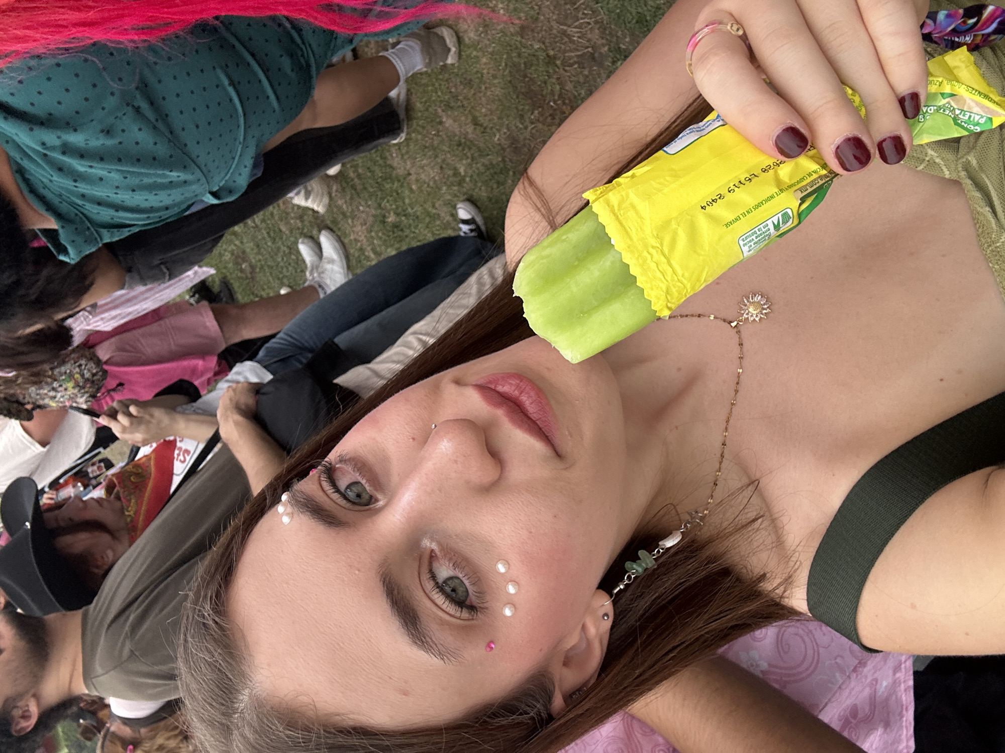 The image shows a woman lying on the ground, holding a green popsicle in a yellow wrapper. The wrapper has text in Spanish, including "PALETA" and "INGREDIENTES: Agua, Azucar". The wrapper also mentions that the popsicle is made in Mexico and has a date code "2628 L6119 2404". The woman has brown hair and green eyes, and is wearing makeup. She has a nose piercing and small pearl-like decorations on her face. She is wearing a necklace with a sunflower pendant. Her nails are painted dark red.
In the background, there are several other people on the ground. Some of them are sitting or lying down. One person has pink hair and another is wearing a black baseball cap.
Based on the language on the popsicle wrapper, this scene is likely taking place in Mexico. Without more specific information, the location in Mexico City cannot be determined.