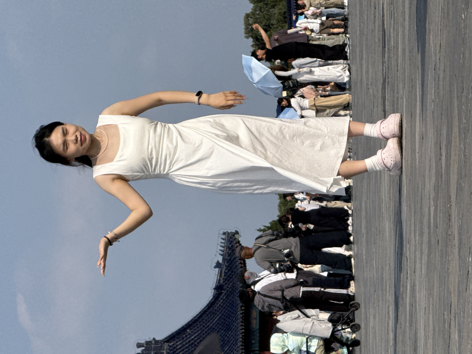 A young woman in a white dress and pink Crocs stands on a wooden boardwalk, holding her left hand open, palm up, as if presenting something. She is in the foreground, facing slightly left, with her eyes squinted. Behind her, a crowd of people mills about. To the left, a traditional Chinese temple with a dark blue tiled roof is visible. The scene appears to be outdoors under a blue sky with some clouds. The woman is wearing a delicate pearl necklace and a pink fitness tracker on her left wrist. In the background, people are seen holding umbrellas, taking photos, and pushing a stroller. The location is Dongcheng District, China.