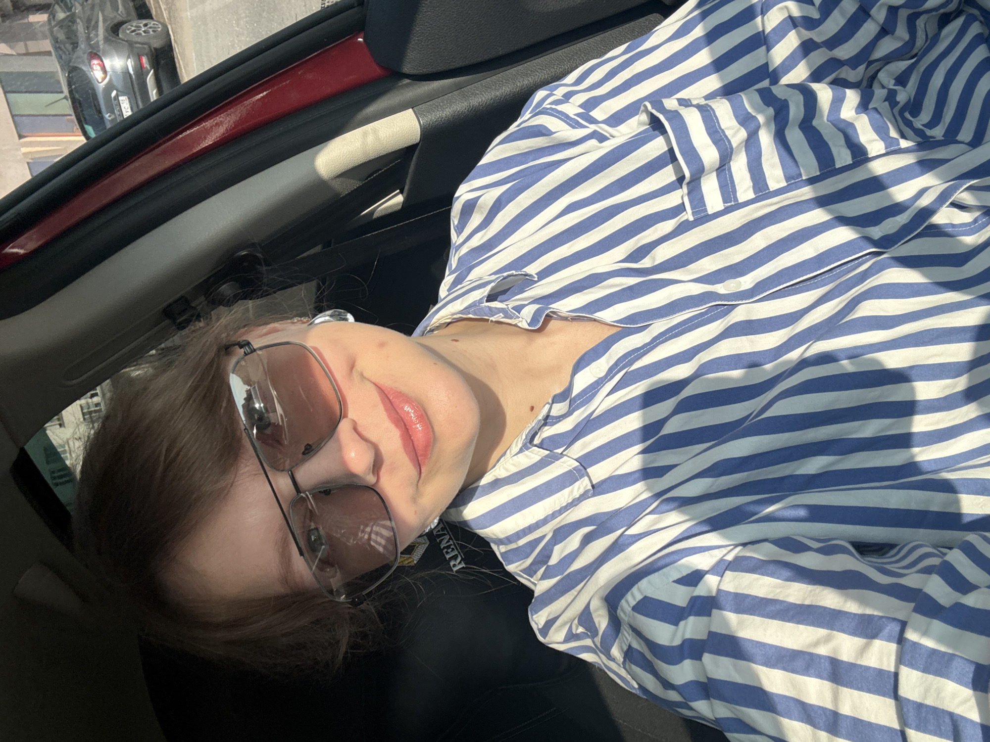 A close-up, selfie-style, medium shot captures a Caucasian woman with light brown hair, wearing large, aviator-style sunglasses, and a blue and white striped button-down shirt. She is seated in the driver's seat of a car, with the car's interior visible in the foreground. The car is a dark red, and a portion of a black leather seat with the word "RENAULT" embossed on it is visible. The woman is smiling with her lips slightly parted, and her makeup includes lip gloss. She is also wearing large, silver hoop earrings. The background shows a street scene in Izhevsk, Russia, with a grey Renault SUV parked on the right side of the image. The lighting suggests it is daytime, with strong sunlight casting shadows.