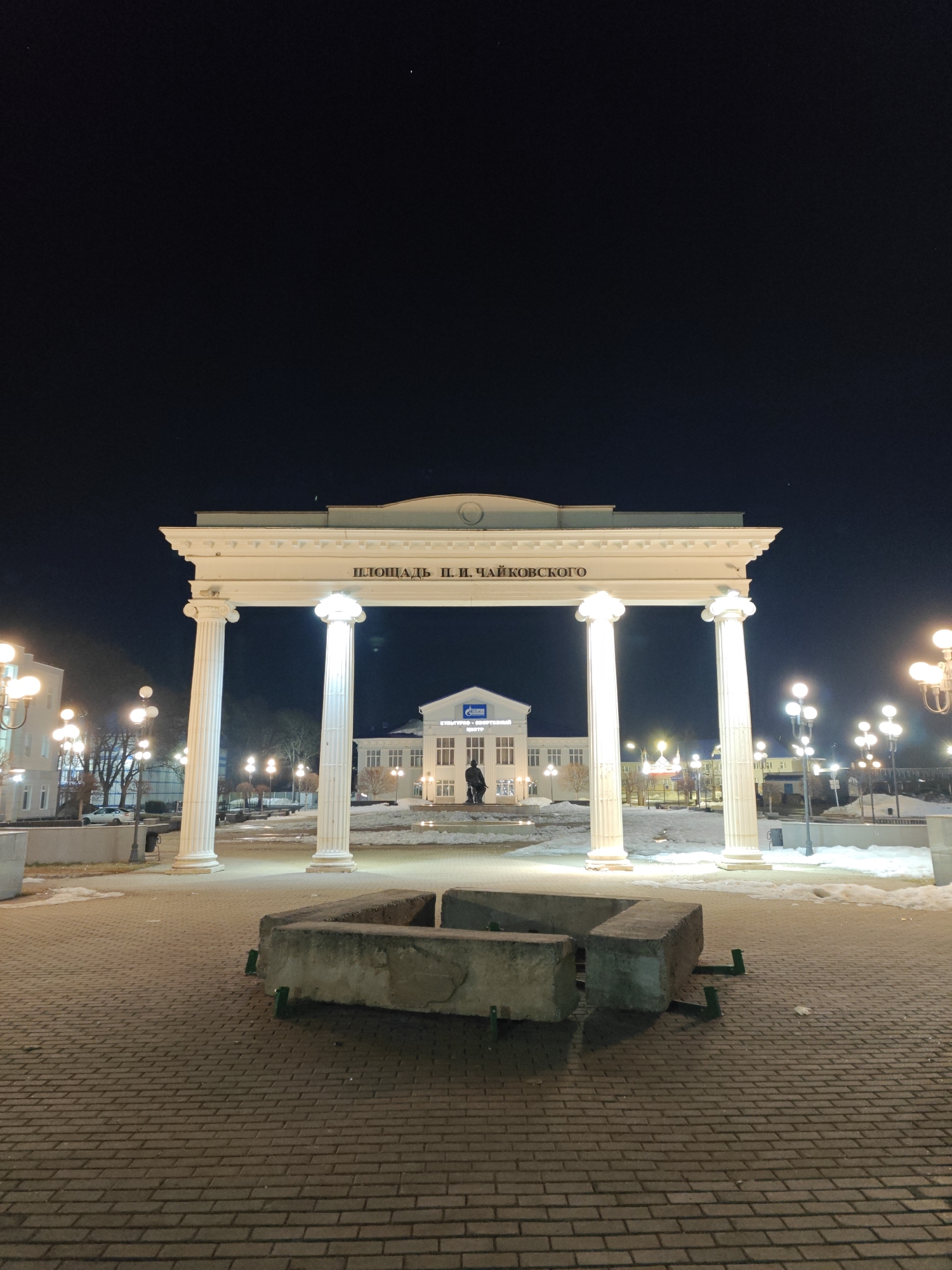 The image depicts a nighttime scene in Chaikovsky, Russia. The foreground shows a paved plaza with a low, rectangular concrete structure in the center. Behind it stands a white neoclassical archway supported by columns, with the inscription "Площадь П. И. Чайковского" (Tchaikovsky Square). In the background, a building with a Gazprom logo can be seen. Street lamps illuminate the area, and patches of snow are visible on the ground. A statue stands in front of the Gazprom building. The sky is dark and clear.