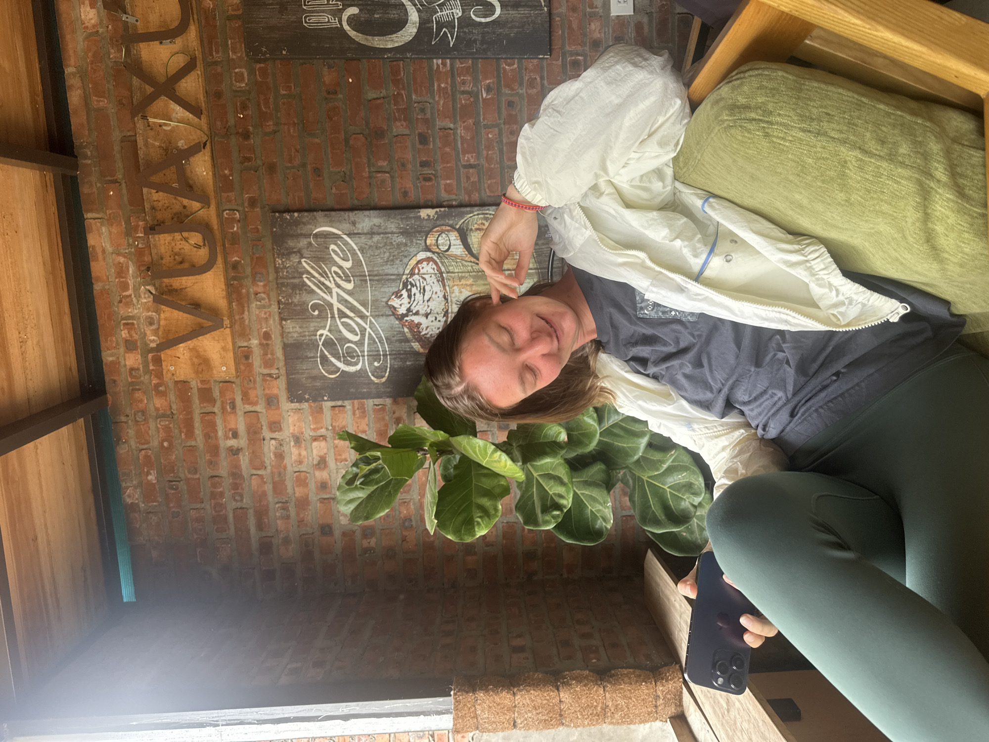 An indoor scene captures a woman relaxing comfortably in a cafe in Mù Cang Chải, Vietnam. She is seated, leaning back against a light green cushion, her eyes closed in a relaxed expression with a gentle smile. Her light brown hair frames her face. She wears a light-colored windbreaker over a dark grey t-shirt and dark green leggings. Her right hand is lightly touching her ear, while her left hand holds a dark smartphone, resting on her leg.
In front of her is a lush, large-leafed green plant, possibly a Fiddle-leaf fig, adding a touch of nature to the interior. The cafe features a rustic aesthetic, with a prominent brick wall in the background. Adorning the wall are distressed wooden signs. One sign clearly displays the word "Coffee" in a stylized white script, accompanied by an image of a coffee cup. Another set of large, dark letters spelling "VUXA" is partially visible on the brick wall to the left, adding to the cafe's unique decor. Wooden furniture elements are also visible, including a frame around the cushion she sits on and a wooden surface to her left. The overall atmosphere suggests a peaceful and cozy daytime setting.