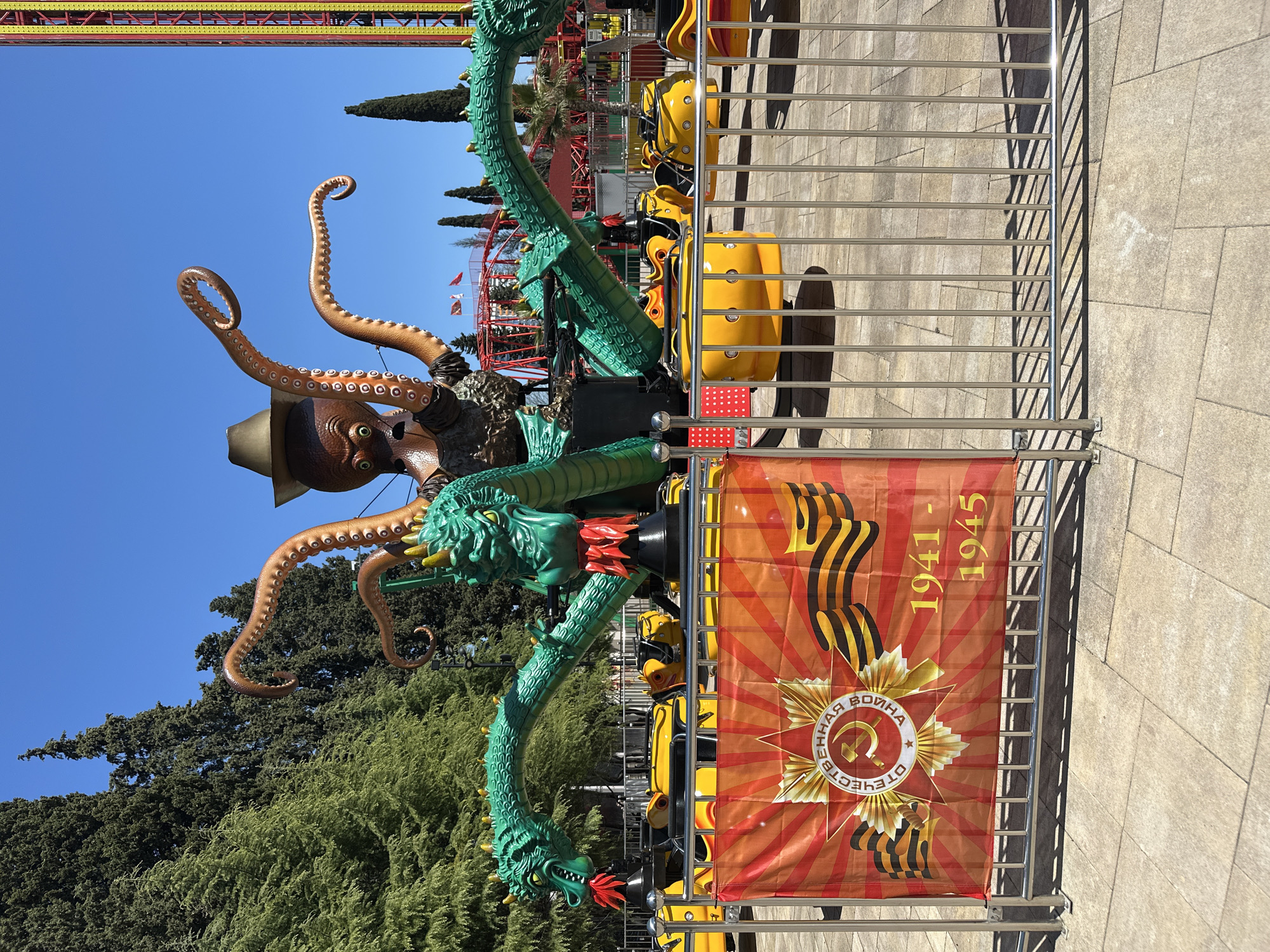 This vibrant image depicts a section of an amusement park in Sochi, Russia, bathed in bright sunlight under a clear blue sky. The central feature is a colorful, elaborate ride. It incorporates a large, brown octopus figure wearing a dark hat, with textured tentacles curling upwards. Coiling around its base and extending into the ride structure are several green dragon figures, adorned with red and yellow accents, one of which appears to be breathing "fire" from its mouth with red and orange elements.
Behind a sturdy silver metal fence, bright yellow ride cars with black seats are visible, suggesting a spinning or swinging attraction, currently stationary. The ground is paved with light-colored, square tiles, upon which sharp, distinct shadows from the fence are cast, indicating a sunny day.
Attached to the metal fence in the foreground is a prominent banner. The banner features a red and orange design with radiating lines and integrates the emblem of the Order of the Patriotic War, characterized by a red star, hammer and sickle, and laurel branches. Text on the banner clearly reads "ОТЕЧЕСТВЕННАЯ ВОЙНА" (Patriotic War) and the dates "1941 - 1945", commemorating the Great Patriotic War.
In the background, lush green trees, likely conifers such as cypress, line the horizon, with glimpses of other amusement park structures, including red and yellow roller coaster tracks, visible further in the distance. No people are discernibly present in the immediate vicinity of the ride.