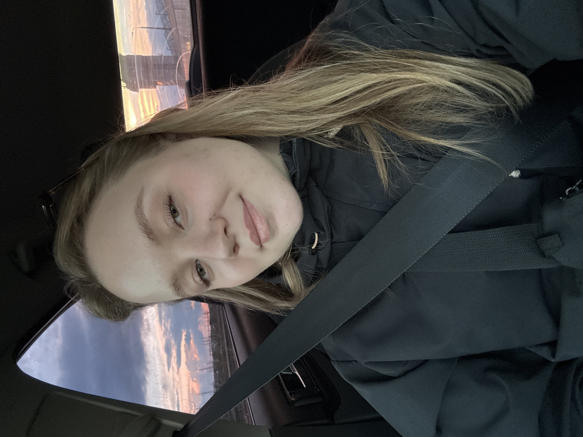 A selfie taken inside a car. A woman with blonde hair is wearing a black jacket and a seatbelt. Outside the car window, there is a view of what appears to be a sunset and a bridge. The photo was taken in the city of Chaikovsky, Russia.