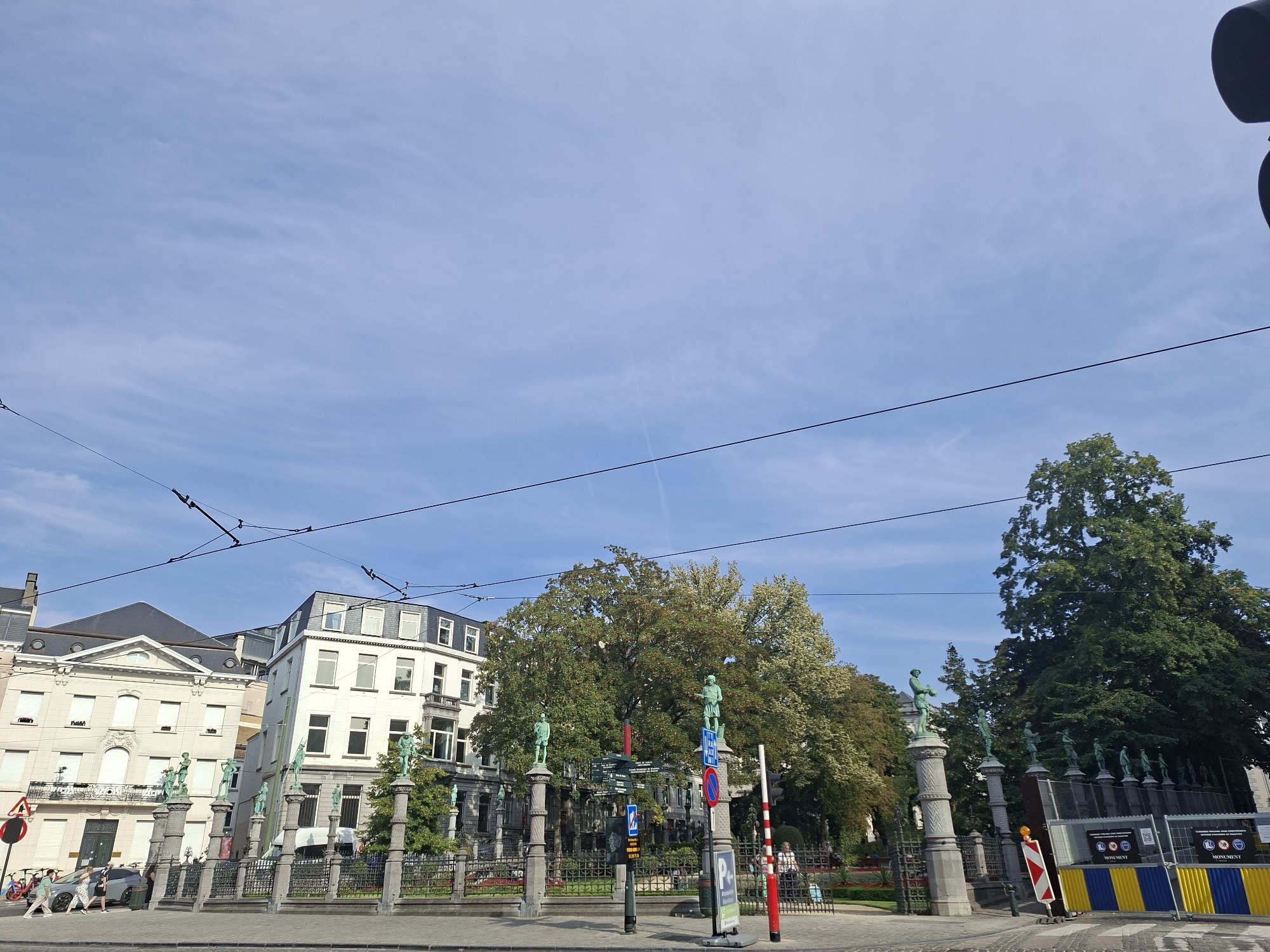 The image captures a view of the Place Saint-Géry in Brussels, Belgium, on a clear day. In the foreground, a street separates the viewer from a park-like area bordered by a black metal fence. Several statues of figures stand atop stone pillars in front of the fence. Behind the fence, trees and buildings are visible. One building, to the left, is light-colored with classical architectural details. To its right, a taller, white building with multiple windows and balconies is positioned at an angle. Further back, a cluster of mature green trees dominates the center of the frame, with other buildings partially obscured by the foliage. Several people are present: two women walk on the sidewalk in the lower left, and others are visible near the fence and in the background. Overhead, a network of electric wires and poles suggests the presence of public transportation, likely trams, which are common in Brussels. Construction barriers with yellow and blue panels are also visible in the lower right section of the image.